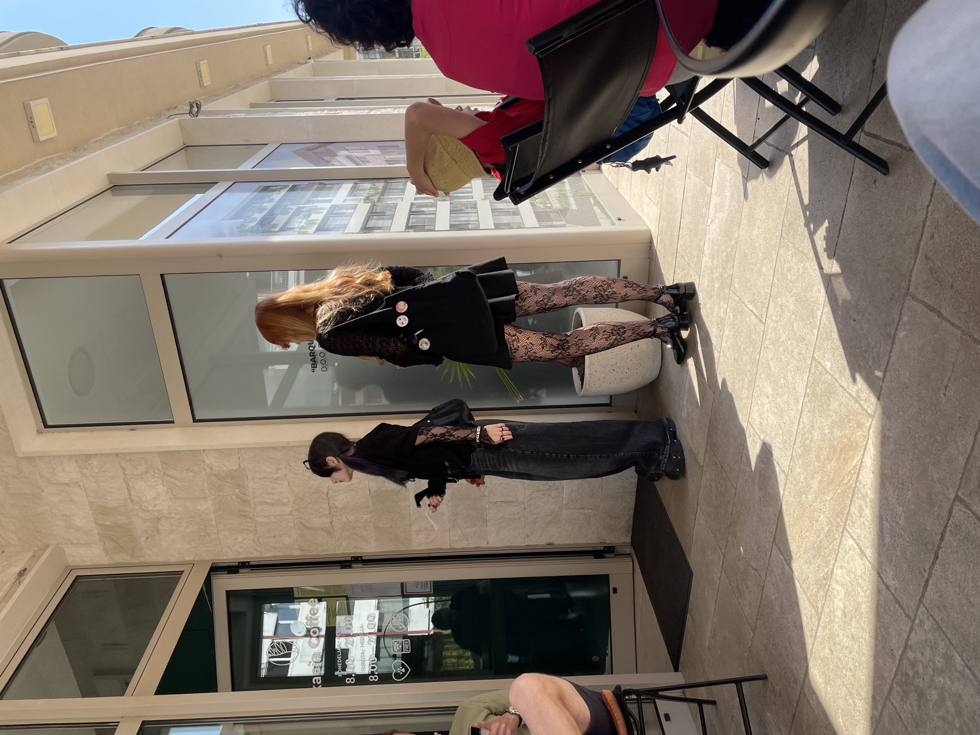 The image depicts an outdoor scene in front of a modern storefront, likely a cafe, under bright sunlight. The ground is paved with light-colored square tiles, and strong, elongated shadows are cast by tables and chairs, indicating either morning or late afternoon.
Several individuals are present. In the foreground, on the right, a person with dark hair wearing a red top is seated at a black metal table. Behind them, further right, another person with a red top and a light-colored hat is also seated. Closer to the building, two people are standing. One individual has long, light brown/auburn hair, wearing a black dress adorned with several pins, patterned lace stockings, and black shoes, standing next to a white cylindrical planter. Adjacent to them, a person with dark hair wears a black long-sleeved top with lace detailing, dark jeans, and dark shoes, appearing to be looking down while standing near the entrance. Partially visible in the bottom left corner, another person in an olive-green top and dark shorts is seated, holding a mobile phone.
The building on the left features large glass windows and an entrance door. Reflections of nearby structures are visible in the windows. Above the storefront, the building facade is light-colored with integrated light fixtures.
Visible text on the glass entrance door includes "Kara Coffee", and "PONEDELJAK - PETAK" (Monday - Friday) with operating hours "8.00 - 21.00". Below that, "SUBOTA - NEDELJA" (Saturday - Sunday) shows "8.00" (implied closing at 21.00). A symbol of a heart with "FREE" is also present. Higher on the window, text reads "BARQU D.O.O." The scene suggests a relaxed atmosphere outside a cafe or shop on a sunny day.