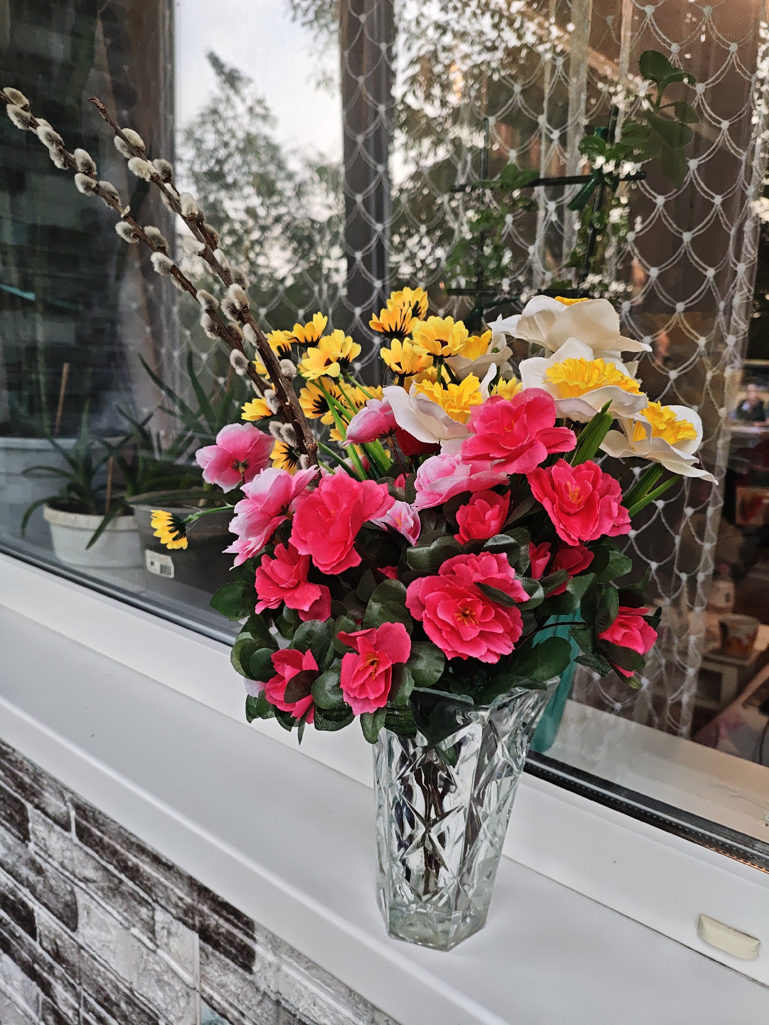 A vase of artificial flowers is placed on a white windowsill. The vase is made of clear glass and has a faceted design. The bouquet consists of pink and red roses, yellow daisies, white daffodils, and pussy willow branches. Behind the flowers, a window with a decorative net curtain is visible, reflecting trees and the sky. The windowsill appears to be part of an apartment or house located in Stavropol, Russia.