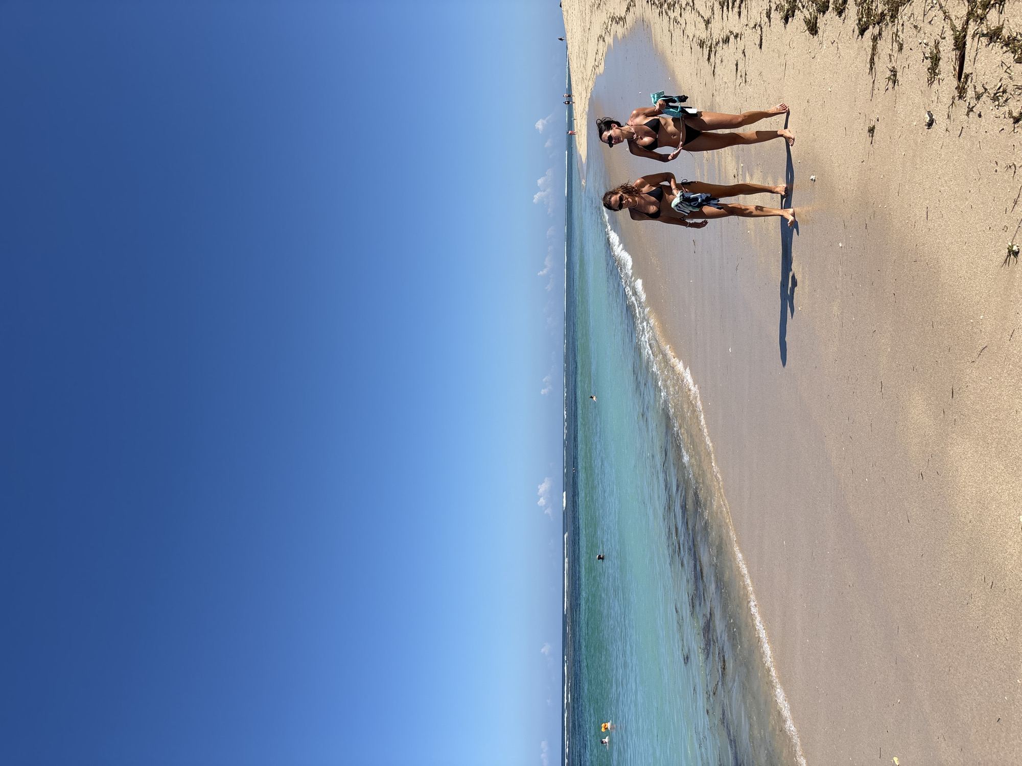 The image shows a beach scene in Nusa Dua, Indonesia. Two women in bikinis stand on the sandy shore near the water's edge. They both wear sunglasses and appear to be holding small objects in their hands. The clear turquoise water laps gently against the beach. The sky above is a cloudless, vibrant blue. The sand is a light golden color, and there is some vegetation towards the edge of the beach. Several people are visible in the ocean. The scene is bright and sunny.