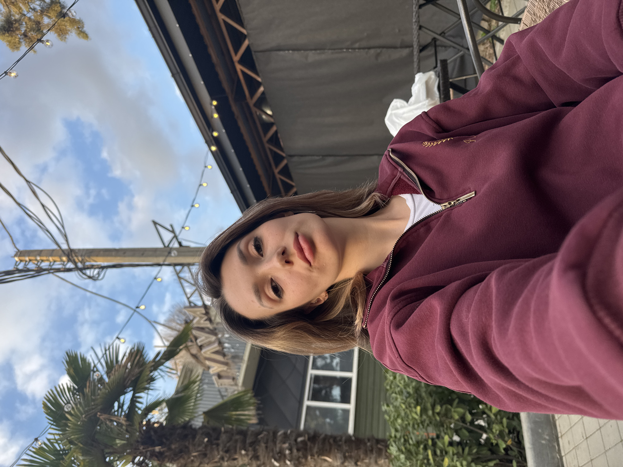 A selfie-style portrait captures a young woman outdoors during the daytime, with the image rotated ninety degrees counter-clockwise. The woman is positioned on the right side of the frame, looking directly at the camera with a neutral expression and slightly parted lips. She has light brown hair and is wearing a maroon or burgundy zippered hoodie over a white top. A subtle gold-colored embroidered design is visible on the left chest area of her hoodie.
The background reveals an outdoor setting under a partly cloudy blue sky. Several palm trees are visible, predominantly on the left side of the frame. A wooden utility pole with multiple wires crosses the middle ground. String lights with warm-toned bulbs are strung across the scene, appearing both in the foreground and receding into the background. Behind the woman and to her right, there's a dark-colored building or structure with windows, and what appears to be an overhead awning or roof. Parts of dark metal outdoor furniture and some white fabric are also present on the right. The overall impression is that of a relaxed outdoor environment, possibly a patio or garden area, during daylight hours. No discernible text is visible.