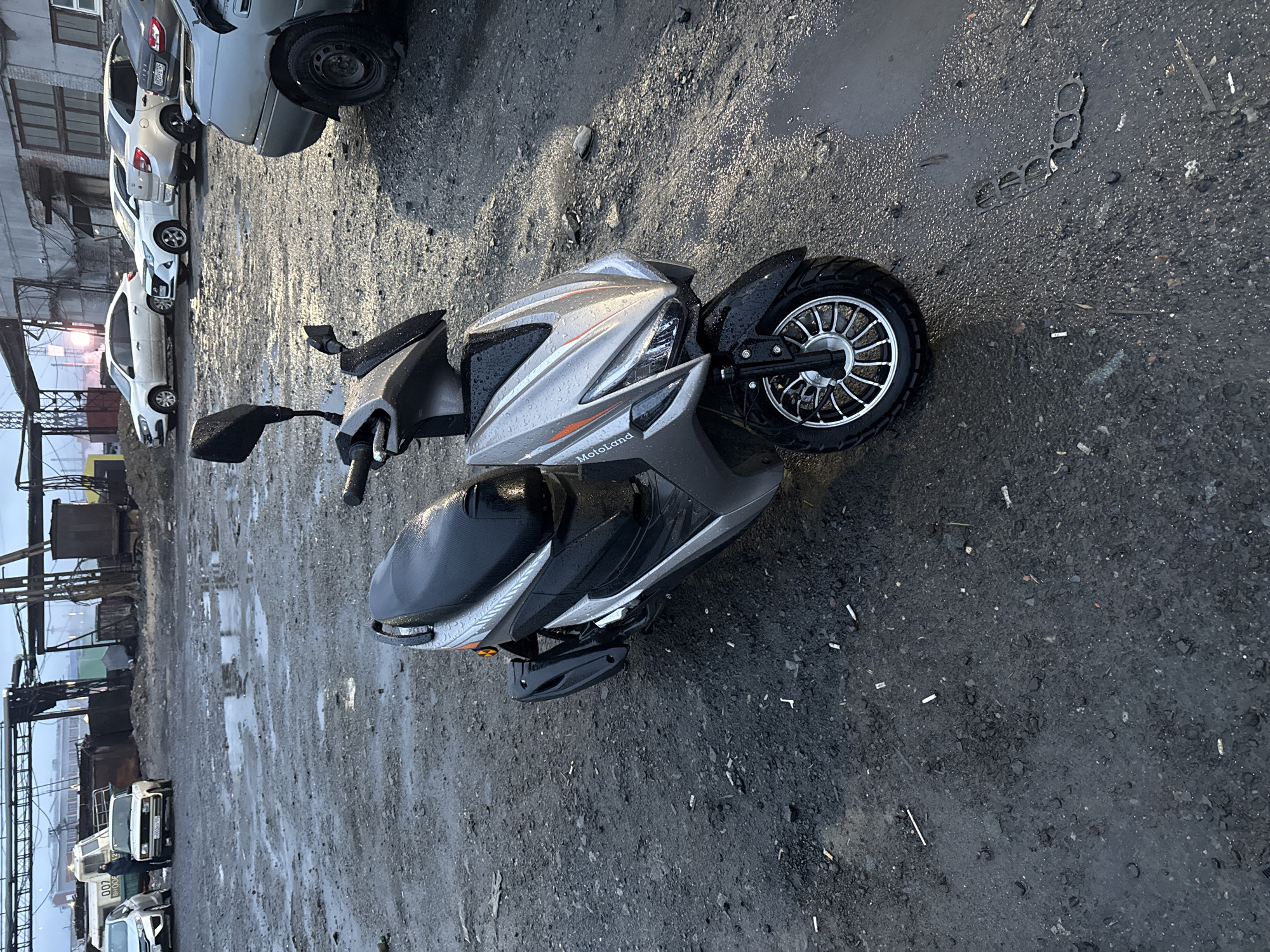The image depicts an outdoor scene in an industrial area of Norilsk, Russia, characterized by a damp, gritty, and uneven ground surface littered with debris, including cigarette butts and what appears to be discarded gasket material. The sky is overcast, and puddles of water indicate recent precipitation. In the foreground, a silver and black electric scooter is parked, its surfaces glistening with water droplets. The scooter has a "Motoland" logo visible on its front panel. In the background, several cars are parked on the wet ground. To the left, a white hatchback and a white sedan are visible, along with a dark-colored utility vehicle. Further in the background, industrial structures and buildings are present, some appearing weathered and industrial. The overall impression is one of a utilitarian, possibly neglected, urban fringe or industrial zone.