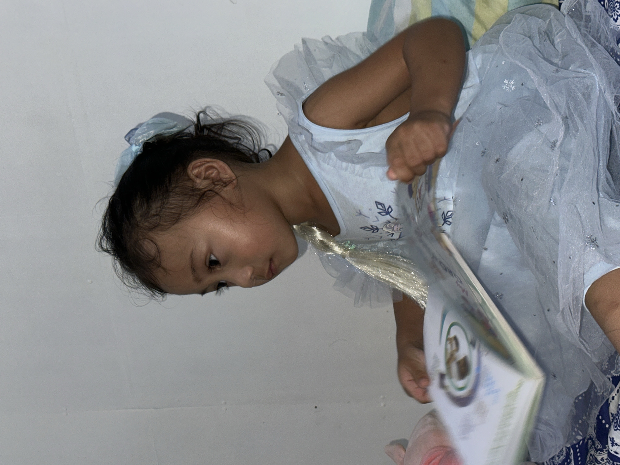 A young girl, appearing to be of Southeast Asian descent, is seated and engrossed in a book. She is wearing a light blue, frilly dress with silver snowflake embellishments, suggesting a themed costume, possibly of a princess. Her dark hair is pulled back with a light blue bow. The girl is looking to her left, intently focused on the open book held in her hands. The book's pages appear to be illustrated. She is positioned on a surface covered with a patterned blue and white fabric, likely a bed or sofa, with a striped green and yellow cushion visible behind her. The background is a plain white wall. The lighting suggests an indoor setting, possibly in the afternoon or evening. No specific location within Davao City, Philippines, can be determined from the visual information provided.