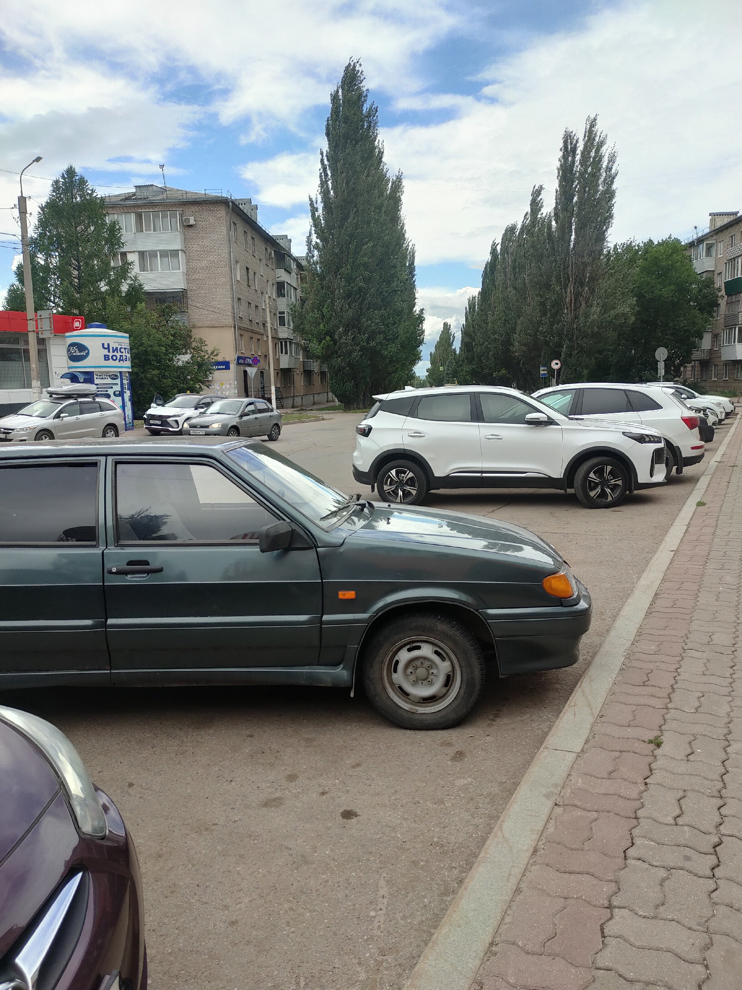 The image captures a street scene in Oktyabrsky, Russia, featuring parked cars and a residential building. In the foreground, the side of a dark green car is visible. Behind it, several white SUVs and a silver car are parked along the street. The buildings in the background are typical of Soviet-era architecture, with multi-story brick structures and balconies. Tall, slender trees line the street, adding a touch of nature to the urban landscape. The sky is partly cloudy, suggesting a mild day. A sign with Russian text is visible on one of the buildings, indicating local businesses or services. The overall impression is a snapshot of everyday life in a Russian city.