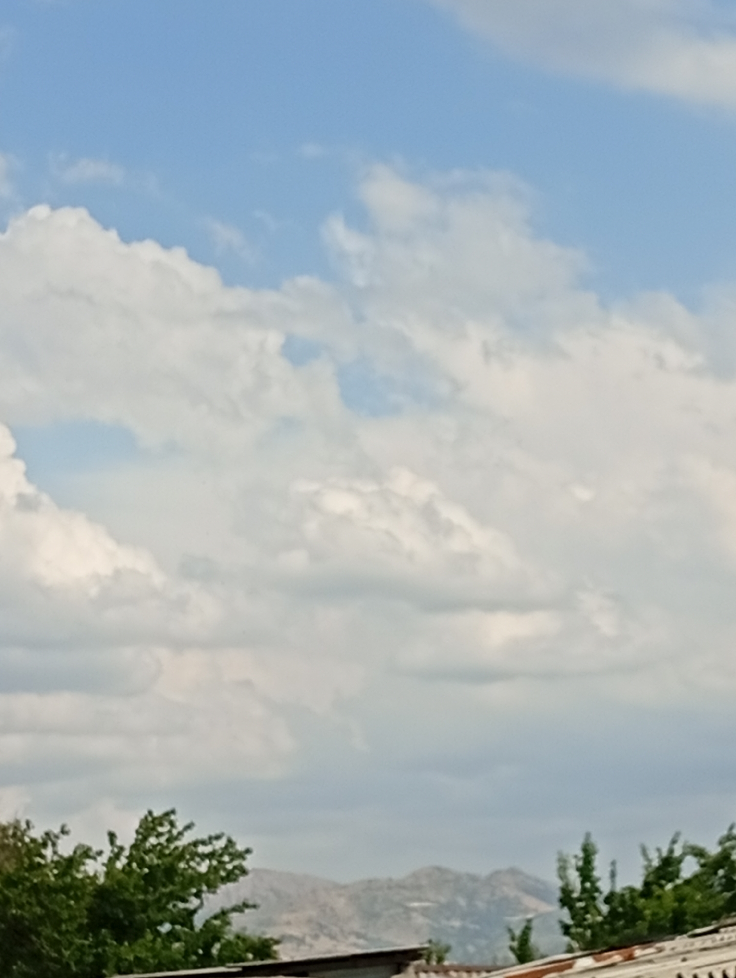 The image depicts a sky with fluffy white clouds against a bright blue sky. Below the clouds, a line of distant, hazy mountains can be seen, some of which are sparsely covered with vegetation. In the foreground, the tops of green trees are visible, along with a portion of a corrugated metal roof. The scene appears to be an outdoor view from a slightly elevated perspective, possibly a rooftop, in the city of Angren, Uzbekistan. The overall impression is of a clear day with a natural landscape backdrop.