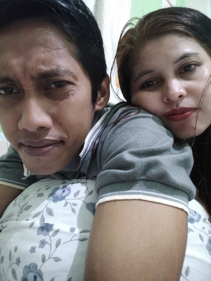 The image displays a close-up indoor portrait featuring two individuals, an adult male and an adult female. The male, positioned on the left, has dark hair and an olive skin tone, looking directly at the camera with a neutral expression. He is wearing a grey polo shirt with ribbed collars and sleeve cuffs, detailed with white and dark stripes. The female, on the right, has dark hair with brown highlights, parted to the side, and is wearing red lipstick. She rests her head gently on the male's right shoulder or upper arm, looking towards the camera with a soft, slightly smiling expression. Her attire appears to be a grey polo shirt, consistent with the male's. The male's right arm is visible in the foreground, positioned over a white pillow adorned with a blue-grey floral pattern. The background is indistinct, showing light-colored walls or curtains, indicating an interior setting. The specific location within the city of Ranugedang, Indonesia, cannot be determined from the visual information presented.