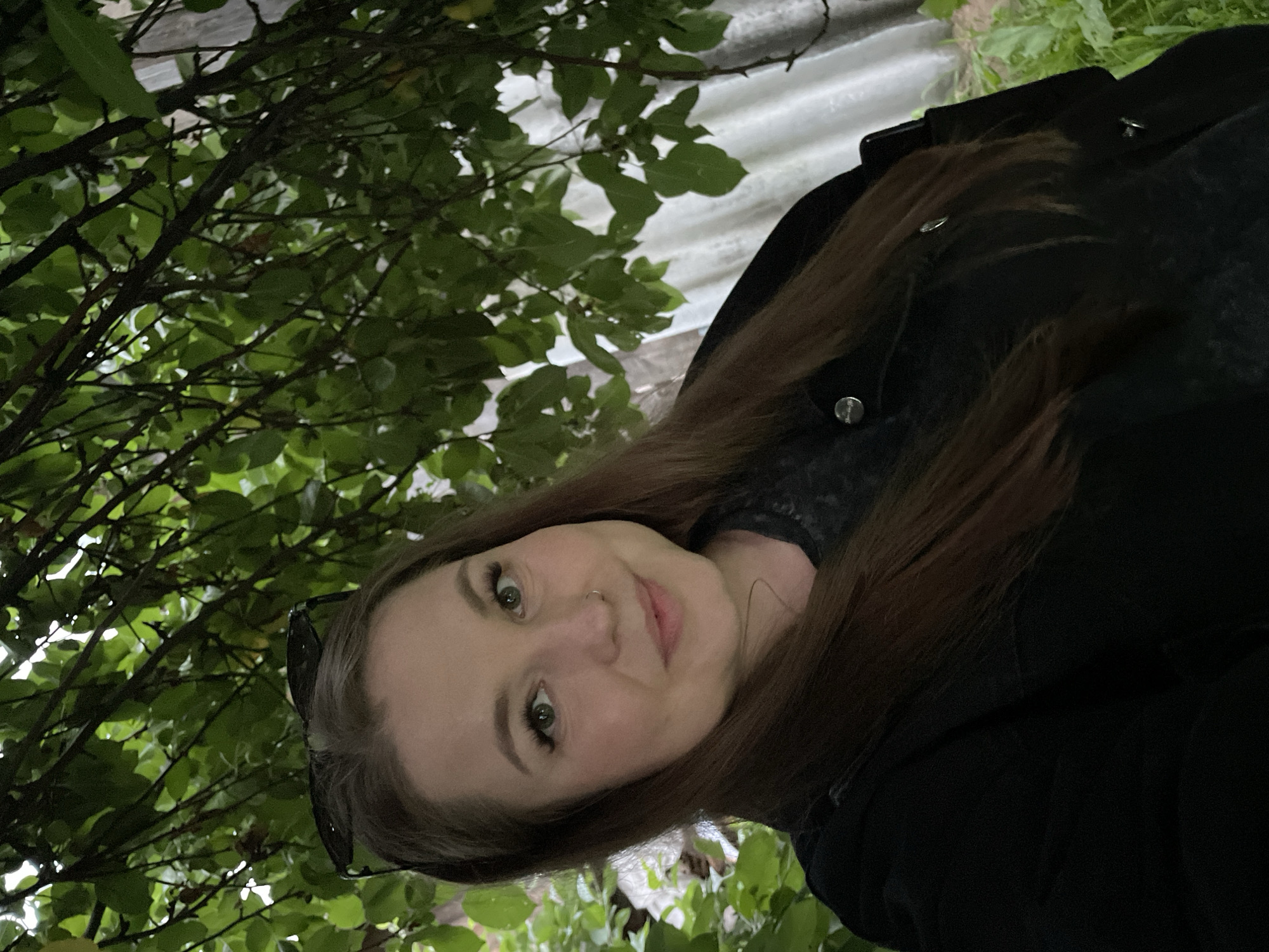 The image is a selfie of a woman with medium-length brown hair, fair skin, and green eyes. She is wearing a black jacket and has sunglasses resting on her head. A small nose ring is visible. She is positioned in front of a backdrop of green leaves from a tree, with branches extending over her head and to the sides. Behind her, there is a corrugated metal structure and some greenery on the ground. The lighting suggests it is dusk or an overcast day. The location is not identifiable within the image.