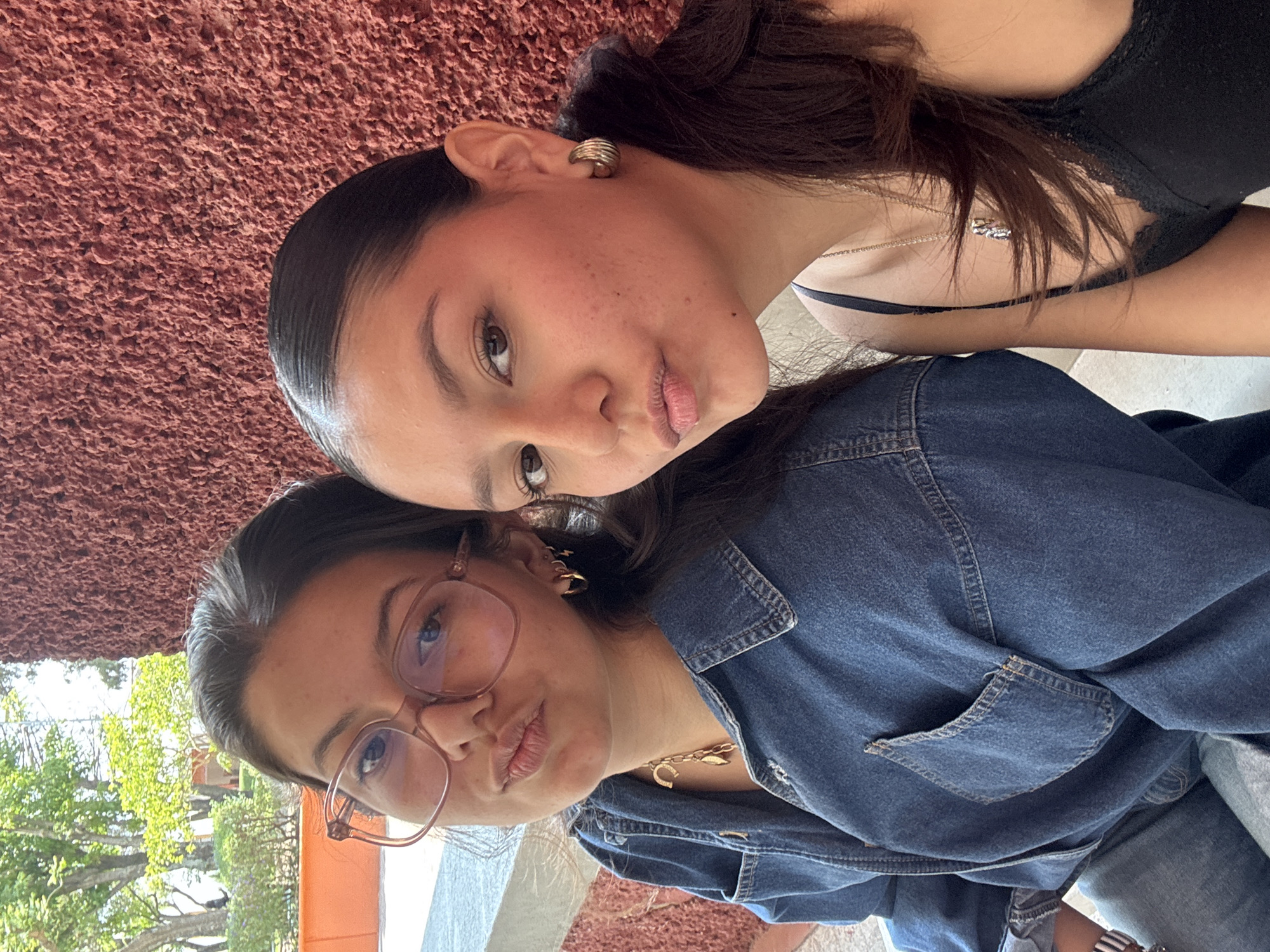 Two young women are posing closely together. One has dark hair pulled back and is wearing a black tank top with a silver necklace and gold hoop earrings. The other woman wears glasses and a denim jacket with a gold necklace. The background appears to be a textured wall with trees visible in the lower left corner, indicating an outdoor setting. This appears to be in San Andrés Cholula, Mexico.