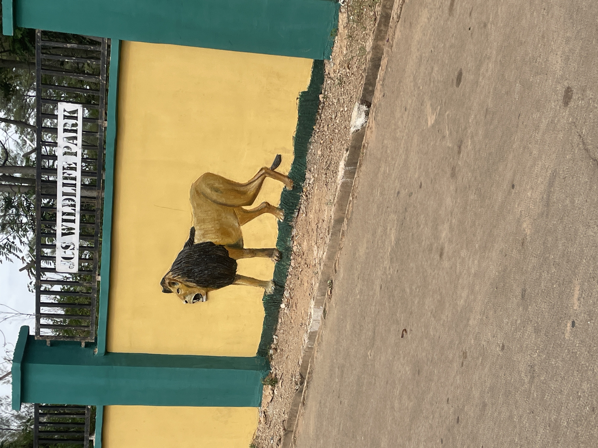 A vertical photograph shows the entrance to a wildlife park in Jos, Nigeria. The entrance features a yellow wall with green trim, adorned with a relief of a lion. The sign reading "Jos Wildlife Park" is vertically oriented in the upper left corner. The foreground is dominated by an asphalt road, indicating vehicle access. The environment suggests a semi-urban or suburban setting. No people are visible in the image.
