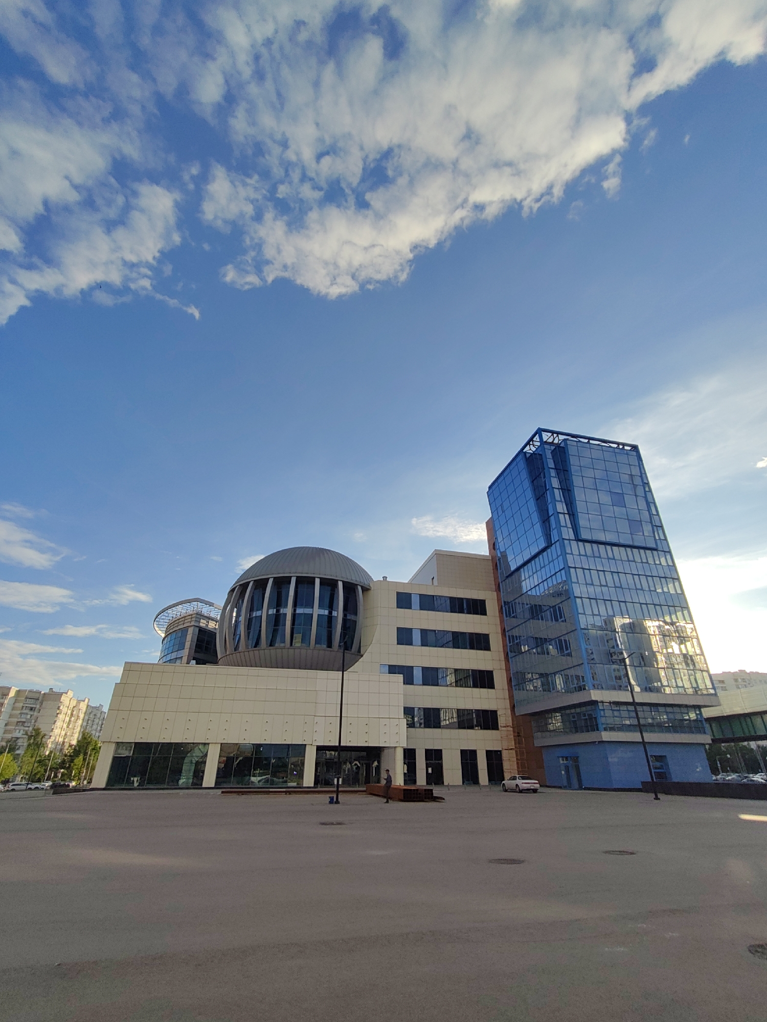The image captures a wide-angle view of a modern architectural complex in Moscow, Russia, under a clear blue sky with scattered white clouds. The scene is static, showcasing the buildings rather than specific activity.
In the foreground, a large, empty paved area, likely a plaza or parking lot, extends across the bottom of the frame. Two primary buildings dominate the midground. To the left and center, a multi-story building features a light-colored facade, possibly stone or composite panels, with large rectangular windows. Its most distinctive feature is a large, ribbed, spherical or dome-shaped structure extending from its upper levels, with vertical window sections. Behind and slightly to the left of this, another, smaller similar dome can be seen.
To the right, an angular, towering structure with a striking blue glass facade rises. Its windows reflect the sky and surrounding environment, including what appears to be sunlight, suggesting the sun is behind or to the side of the viewer. The lower levels of this blue building also feature blue panels.
In the distance to the far left, other residential buildings and trees are visible, indicating an urban setting. A few cars are parked in the distance, including a white car near the base of the buildings. A couple of individuals can be faintly discerned near the entrances or walking across the paved area, but they are not prominent subjects.
The time of day appears to be daylight, possibly morning or late afternoon, given the bright, evenly lit sky and the presence of some soft shadows on the ground. The weather is fair, with wispy and cumulus clouds adding texture to the expansive blue sky. There is no discernible text visible in the image.