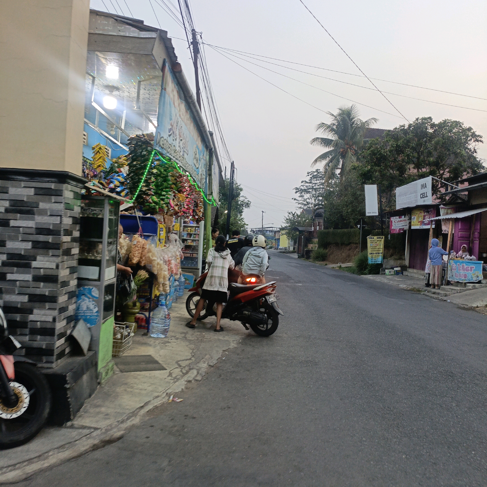 The image depicts a street scene in Salatiga, Indonesia, likely during daylight hours given the ambient light. On the left side of the street, a small shop is visible, displaying various goods for sale, including snacks in colorful packaging, bottled water, and what appear to be bags of chips. The shop features a light blue awning and is adorned with green string lights. Adjacent to the shop, a motorcycle is parked, and two individuals are standing near it, one appearing to be a customer and the other possibly the shopkeeper or another customer.
Further down the street, on the right side, another establishment is present, identified by signage that includes "IMA CELL," suggesting it is a mobile phone shop. Two women are visible near this establishment, one interacting with a service counter or display. The street itself is paved and appears to be a moderately trafficked local road, with overhead power lines visible. Trees, including a palm tree, and some buildings are also present in the background, indicating a semi-urban or suburban setting. The overall impression is of a typical commercial area in a town or city in Indonesia.