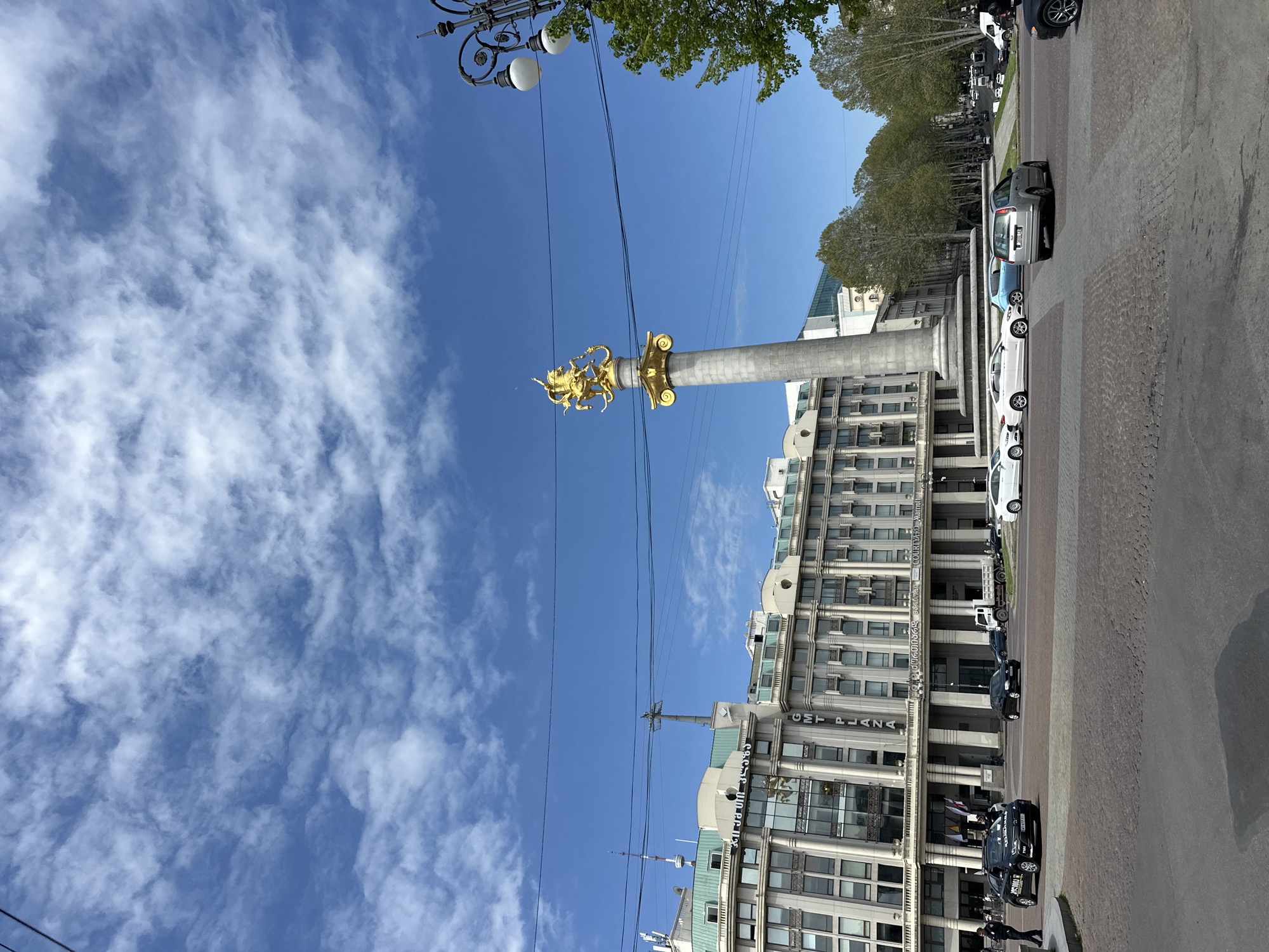 This image captures a vibrant daytime scene in a city square in Tbilisi, Georgia, likely Freedom Square, given the central monument. The photograph is oriented vertically but depicts a horizontal landscape.
The prominent subject is a tall, light-colored stone column, possibly granite or marble, crowned with an ornate golden statue. The statue depicts a powerful figure, commonly identified as Saint George slaying a dragon, characteristic of Georgian iconography. The base of the column features decorative golden elements.
Surrounding the square are multi-story buildings under a bright blue sky with scattered, wispy white clouds. One building on the right (in corrected orientation) clearly displays "CMT PLAZA" in large letters on its facade, with "COURTYARD MARRIOTT" partially visible above it, further confirming the location in Tbilisi. Other buildings with similar architectural styles, featuring numerous windows and structural details, line the background.
The ground consists of a mix of paved areas, some appearing as cobblestones, and asphalt roads. Numerous cars are parked along the street and some are visible in motion, suggesting an active urban environment. Trees with green foliage are present, framing parts of the scene, particularly on the left side of the column. Decorative street lamps with multiple globes and overhead power lines traverse the upper portion of the image. A few indistinct figures of people can be seen near the buildings and vehicles.
The lighting indicates a clear, sunny day, with the sun casting distinct shadows. The overall impression is that of a bustling city center, anchored by a significant national monument.