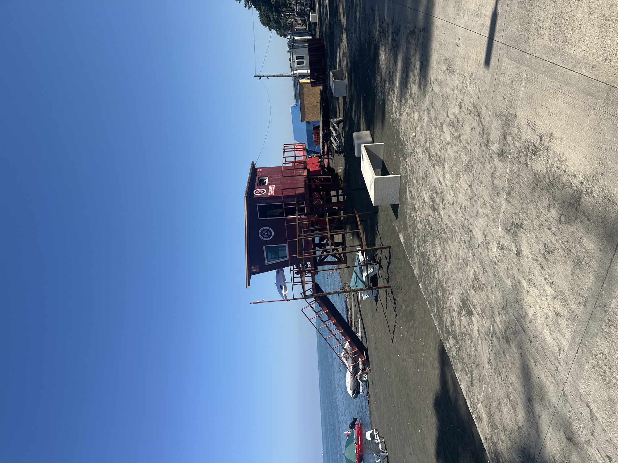 The image depicts a sunny day at a beach in Ureki, Georgia. In the foreground, a concrete promenade is visible with shadows cast by unseen objects. Beyond the promenade, dark sand covers the beach, on which several white benches and loungers are placed. To the left, a red emergency boat with a white canopy and the word "EMERGENCY" is docked near the water's edge. A trailer with a covered boat sits on the sand further back. The central focus of the image is a red lifeguard tower with a ramp leading up to it. The tower has a sign with "112" and other emblems. The calm blue sea stretches out to the horizon, meeting a clear blue sky. In the background, trees and several temporary structures are lined up along the shore. No people are clearly visible in the scene.
