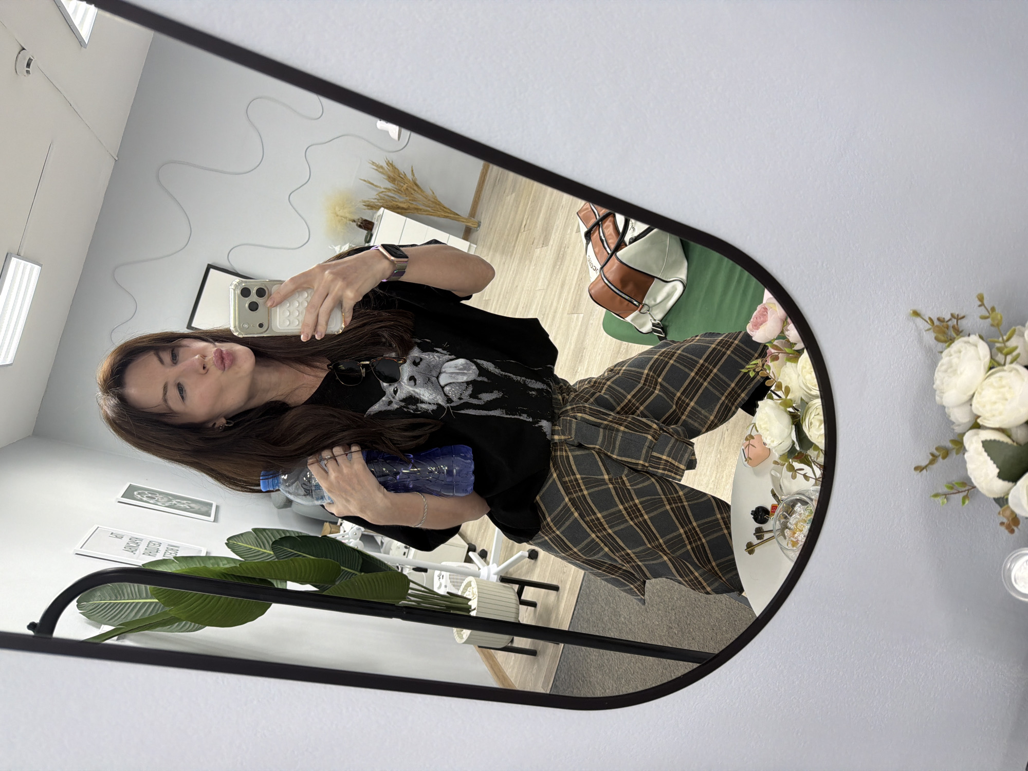 A person is taking a selfie in a mirror inside a room. The individual is holding a water bottle in one hand and a phone in the other, posing with pursed lips. They are wearing a black t-shirt with a graphic design and a plaid shirt tied around their waist. The room has light gray walls with various decorations, including framed pictures, a tall plant, and floral arrangements. Furniture and other items like a white table, a bag on a green chair, and wheat stalks are also visible. The location appears to be indoors, possibly in a shop or studio in Perm, Russia.