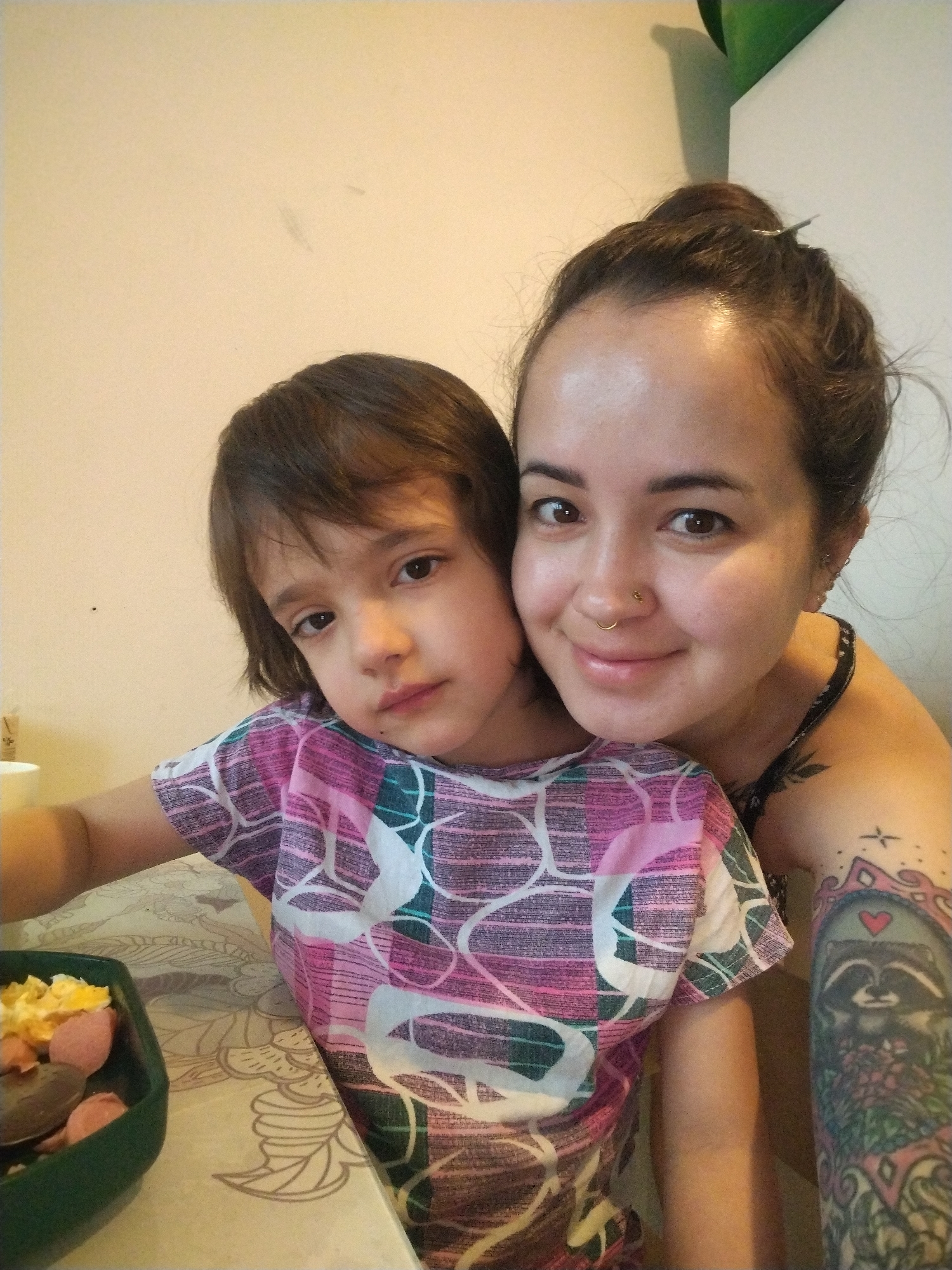 The image captures a close-up portrait of a woman and a child, likely indoors in Noviy, Russia, during what appears to be a casual mealtime. The woman is positioned behind and slightly to the right of the child, looking directly at the camera with a warm smile. She has brown hair pulled back, brown eyes, and notable facial piercings including a nose stud and a nose hoop, along with multiple ear piercings. She is wearing a dark, possibly patterned, strap top, revealing a heavily tattooed left arm. A prominent, colorful tattoo on her forearm features a raccoon with a red heart above its head, framed by intricate designs. Other smaller tattoos are visible on her shoulder.
The child is in the foreground on the left, with shoulder-length dark brown hair and brown eyes, looking towards the viewer with a calm, neutral expression. They are wearing a vibrant pink, white, and teal patterned t-shirt.
In the bottom left of the frame, a green rectangular dish containing scrambled eggs and cut-up hot dogs or sausages is visible, with a spoon in it, suggesting a meal. The subjects are set against a plain, light-colored wall. A light-colored table with a floral or leafy patterned tablecloth is in the foreground. The overall scene conveys a domestic and intimate moment. There is no visible text in the image.