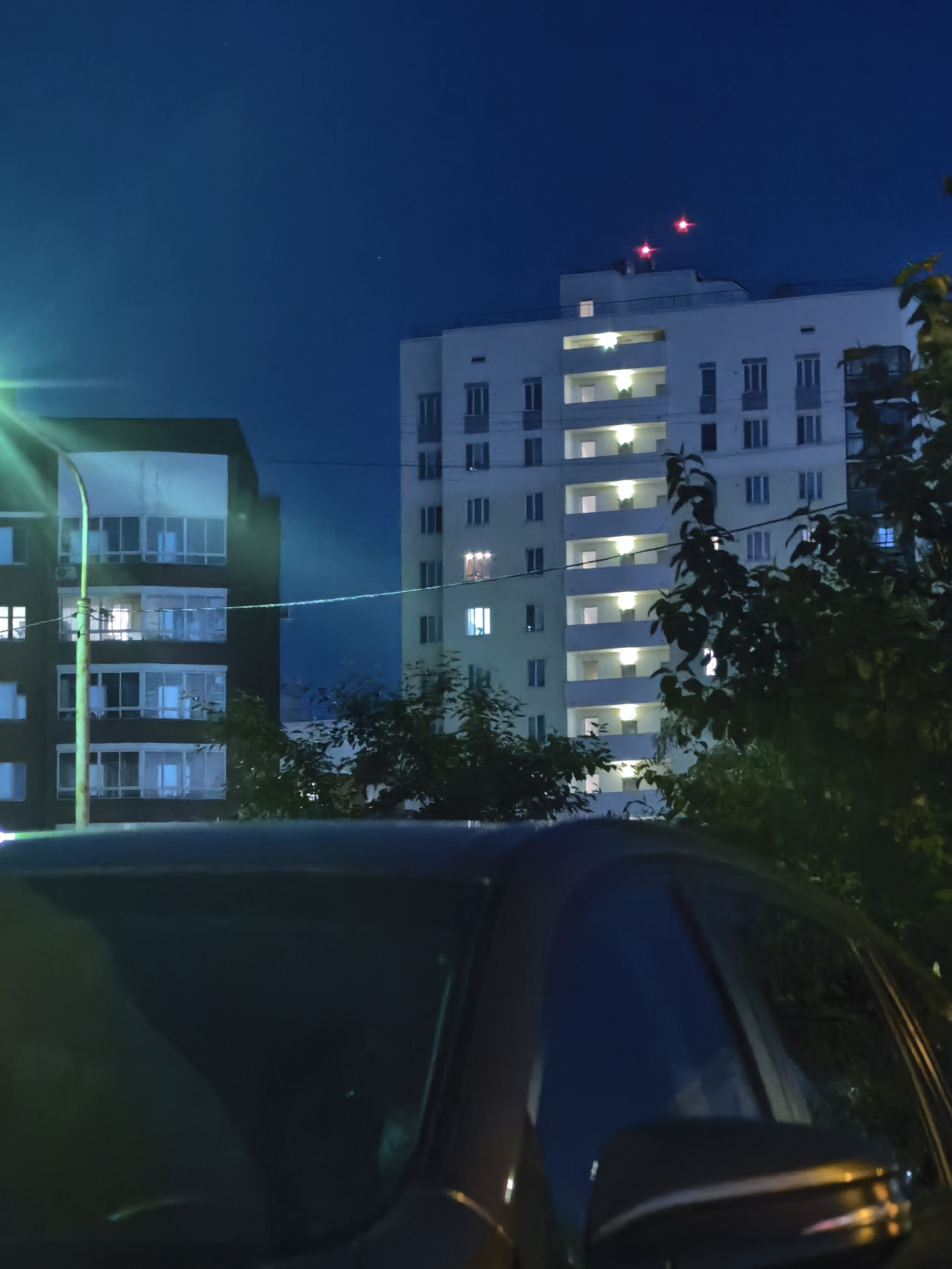 The image depicts an urban scene at night, featuring two residential buildings and the foreground of a car. The building on the left is dark and appears to have balconies and windows illuminated by interior lights. A street lamp with a green light is visible to the left, casting a glow. The building on the right is lighter in color and taller, with multiple floors and windows. Several windows on this building are lit, revealing internal spaces. On the rooftop of the lighter building, two red lights are blinking. Trees with dark foliage are present between and in front of the buildings, obscuring parts of the structures and the sky. The sky is a deep blue, indicating nighttime, and a few faint stars or distant lights are discernible. The car in the foreground is dark and only its front section, including the hood and windshield, is visible. The windshield reflects the surrounding environment. The scene is set in an urban environment in Unknown, Unknown.