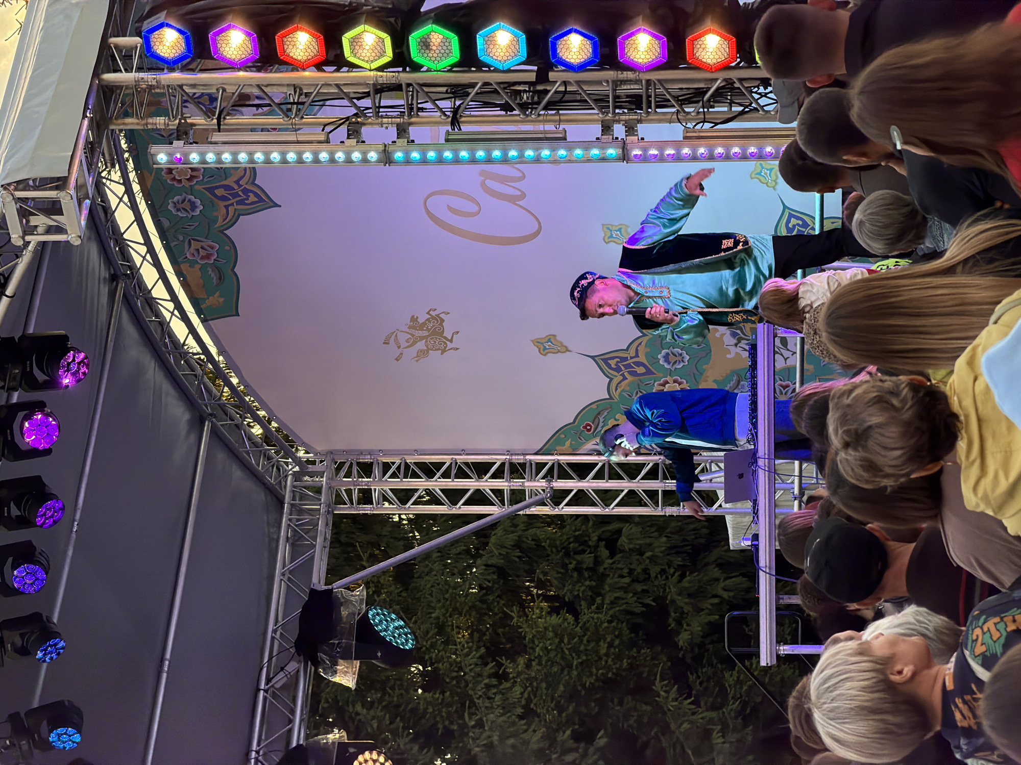 An outdoor stage event is captured from behind a crowd of spectators. Two men are on stage. The man on the left is wearing headphones and a blue jacket, and appears to be operating DJ equipment. The man on the right is wearing a traditional-style turquoise garment with dark trim and a cap, and is holding a microphone and gesturing. The stage has a large white backdrop with a decorative border and lettering. A series of colorful hexagonal stage lights are mounted on a metal truss to the right of the stage. In the background, trees are visible. The audience members in the foreground have their backs to the camera, and are watching the performance.