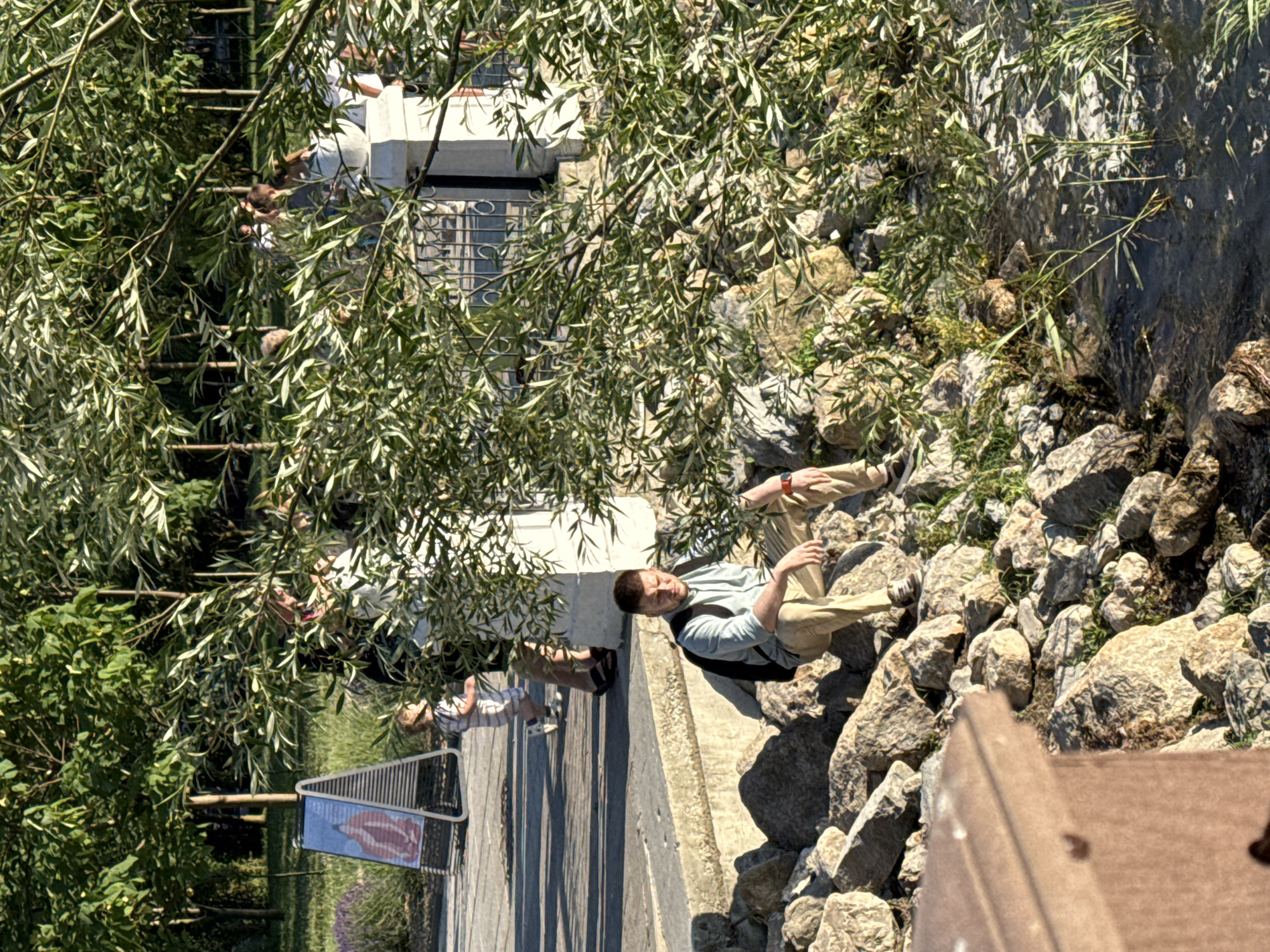 A young man with a backpack is sitting on rocks by a body of water. He is wearing a light blue short-sleeved shirt and tan pants. He is looking down. In the background, several people are standing near a fence and a decorative pillar. A child in a striped outfit is walking on a paved path. There is a sign on an easel in the background. The scene appears to be a park or waterfront area in Abrau-Dyurso, Russia, with trees and vegetation surrounding the area. The lighting suggests it is daytime, likely late afternoon due to the long shadows.