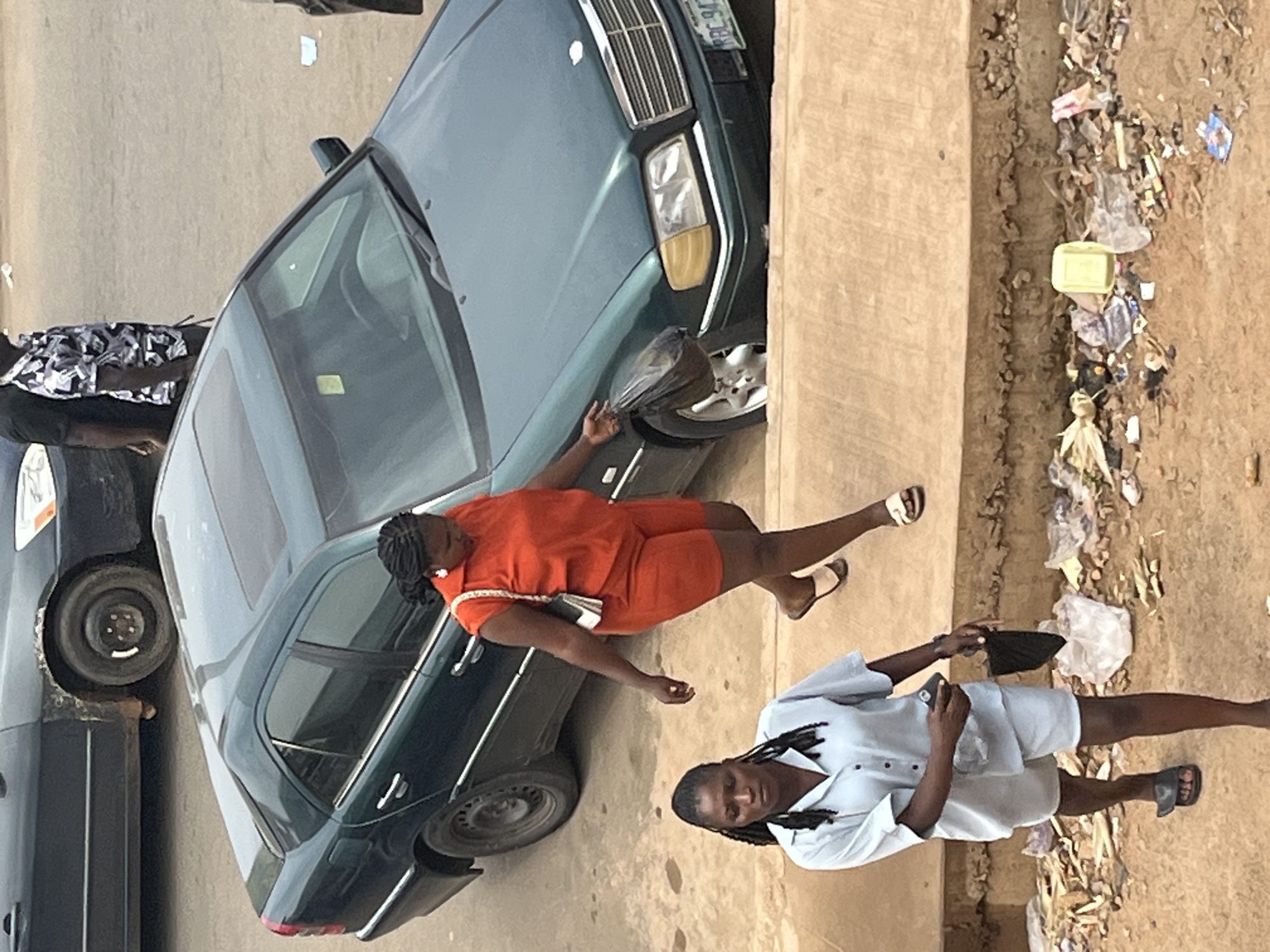 In Jos, Nigeria, a medium shot captures two women navigating a sunlit street. The foreground shows a dirt path littered with refuse, including plastic bags, wrappers, and debris. A concrete embankment lines the path. A woman in a light blue button-up shirt and shorts walks forward, holding a black bag and a phone. Behind her, a second woman in an orange top and shorts walks with a white purse slung over her shoulder and carries a black plastic bag. A dark green Mercedes-Benz sedan is parked in the background, its sunroof visible. Another dark-colored car is partially visible to the left. The scene suggests an urban environment with evidence of street litter and pedestrian activity.