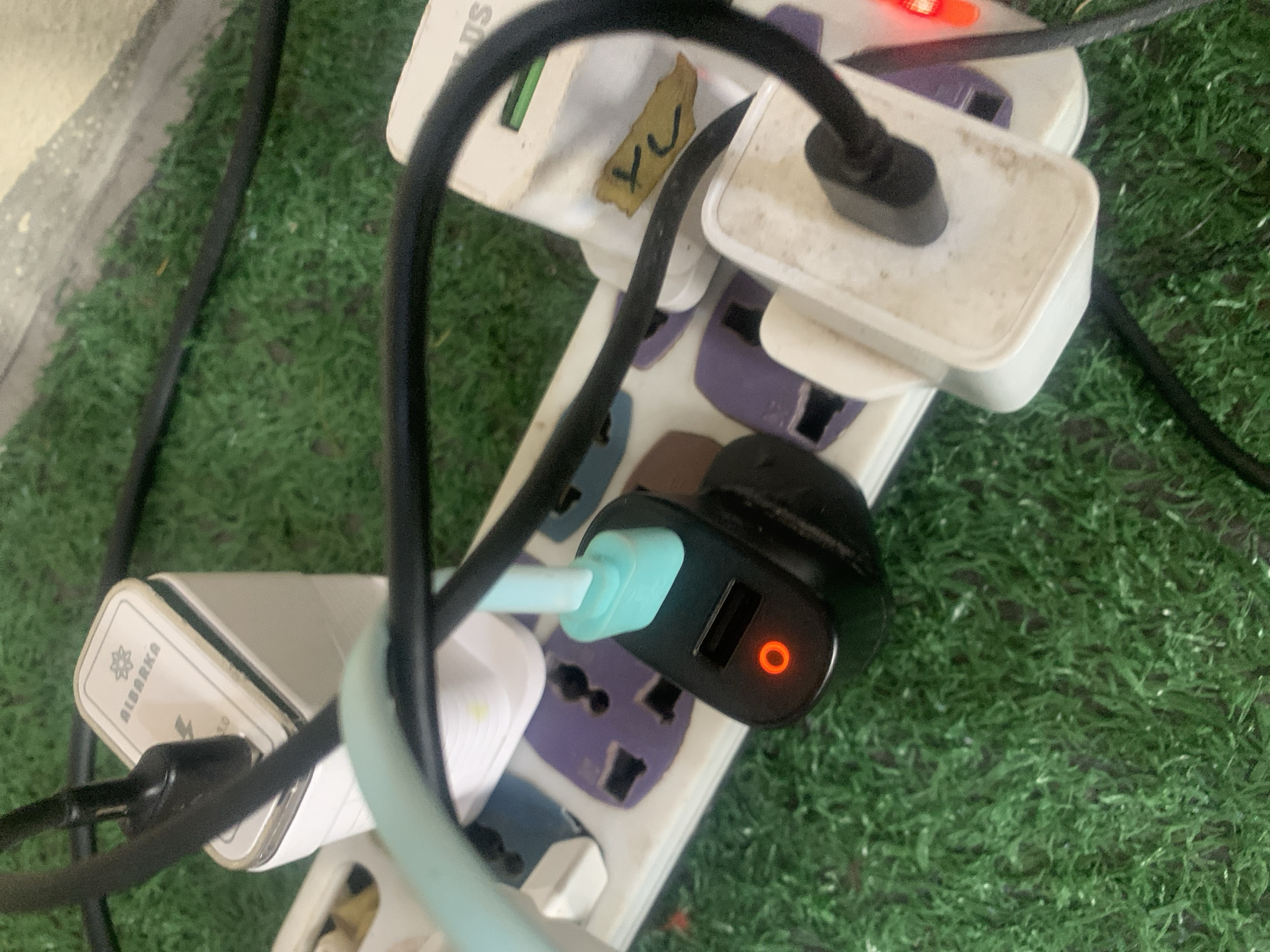 The image shows a close-up, low-angle view of a power strip plugged into an electrical outlet, with several charging adapters and cables connected. The power strip is white with purple and white universal outlets. A black charging adapter with a glowing red indicator light is plugged into one of the outlets, with a light blue USB cable connected to it. Another white charging adapter, labeled "ALBARKA," is plugged into an adjacent outlet. Black cables snake across the scene. The power strip and adapters are situated on a surface covered with artificial grass, suggesting an indoor or possibly outdoor setting not typically associated with Jauro Yinu, Nigeria, as such artificial turf is uncommon in public spaces or common household environments there. No people are visible. The lighting is dim, and the focus is primarily on the electronic devices and their connections.