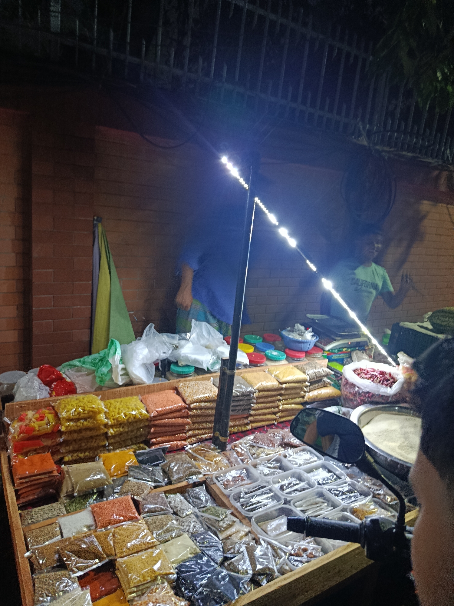 The image shows a roadside spice and grain stall, likely in the city of Dhaka, Bangladesh. The stall is illuminated by a string of LED lights. Various spices and grains are neatly arranged in small plastic bags and containers on a wooden platform. Two men are visible behind the stall, one wearing a green shirt and waving, and the other blurred behind him. A motorbike is parked in front of the stall. The background features a brick wall and a metal fence.