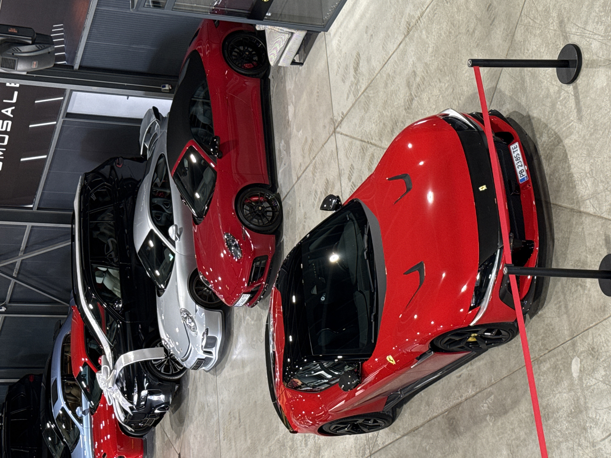 The image shows a car dealership in Plovdiv, Bulgaria, displaying several high-end sports cars. The foreground features a bright red Ferrari, cordoned off by a red barrier tape attached to a black metal pole. Further back, there are other cars in a row, including a red convertible, a silver car, and a black car adorned with a large white ribbon. The dealership has a contemporary design with a glossy floor, likely tile, and a ceiling with visible ductwork. A banner with the text "OMOSALE" is partially visible in the upper left. The license plate "PB 2365 TE" is seen on the red Ferrari.