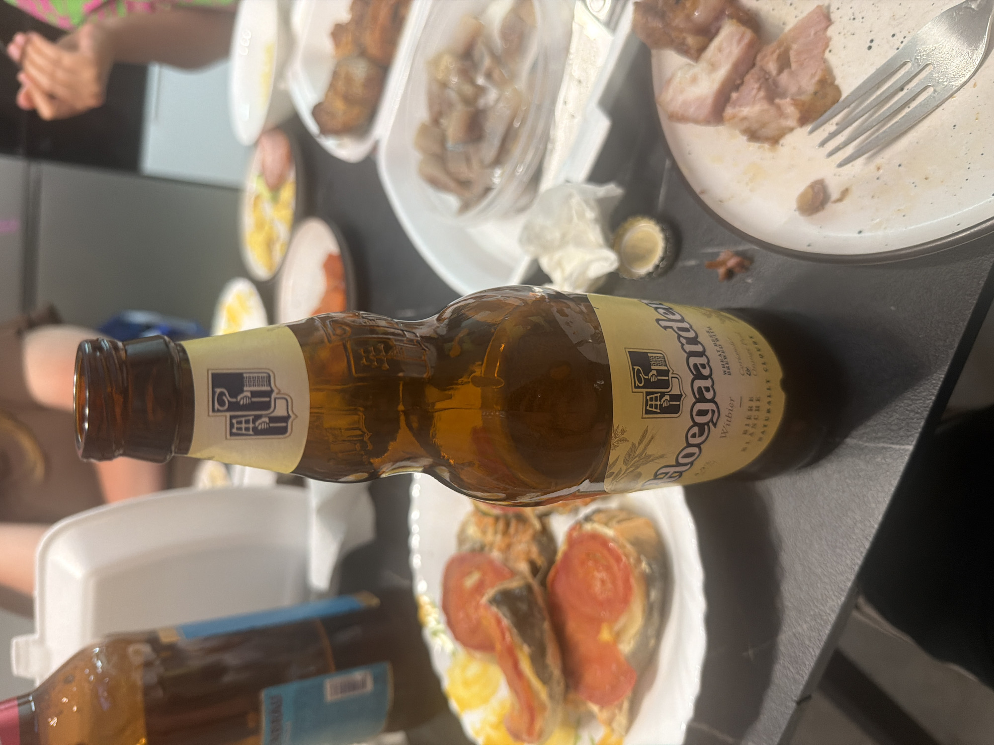 The image depicts a table setting with food and drinks, likely in a casual dining environment. In the foreground, a bottle of Hoegaarden Witbier is prominently displayed. Adjacent to the beer bottle, a bottle cap rests on the table. Several plates of food are visible, including what appears to be open-faced sandwiches with tomato slices, a container of chopped meat or fish, and a plate with pieces of cooked meat and a fork. In the background, other food items and drinks are partially visible, along with what seems to be people's hands and torsos, suggesting a gathering or meal. The overall setting appears to be indoors, on a dark-colored table. There is no information in the image to suggest a location in Novorossiysk, Russia.