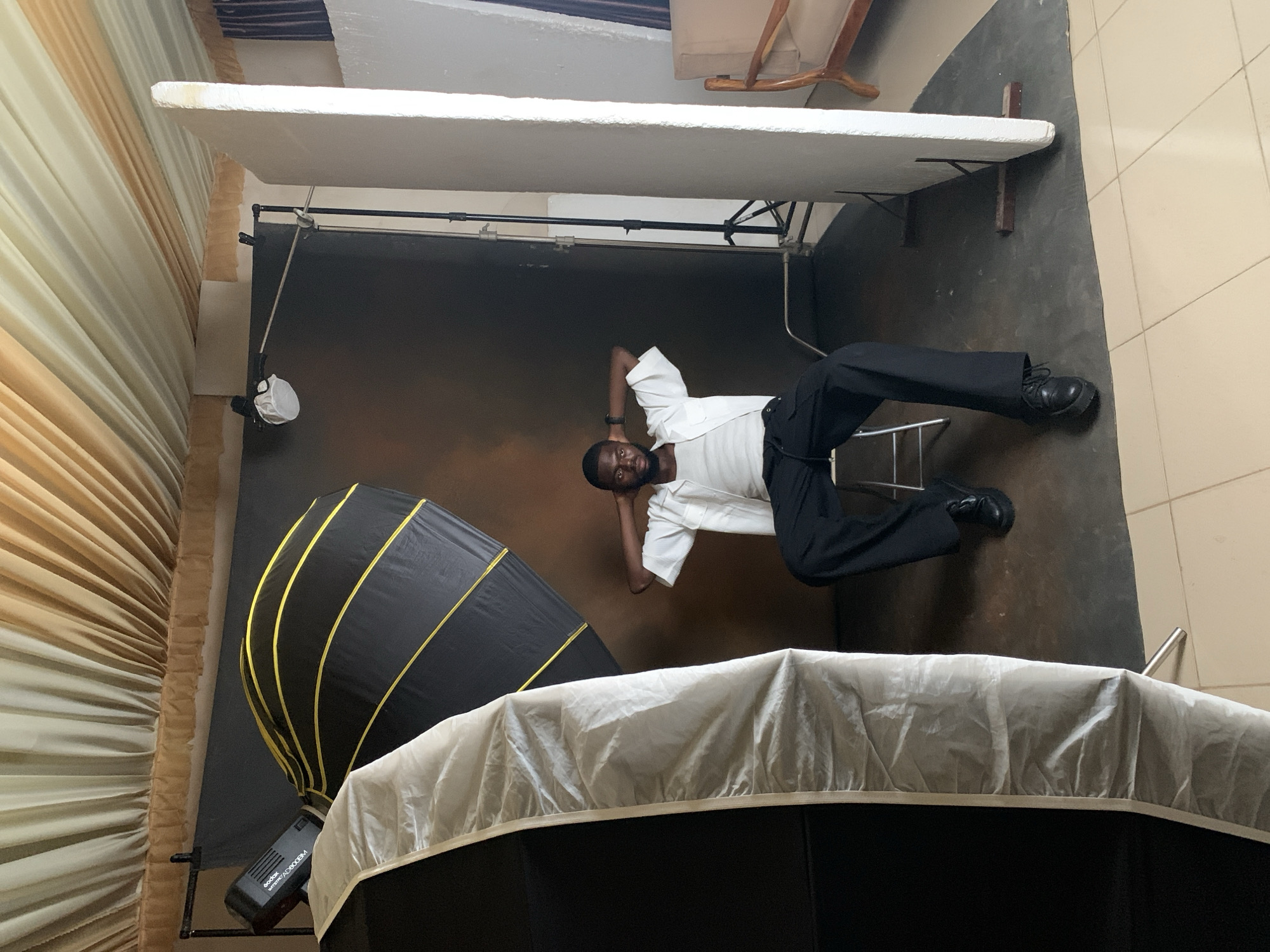 The image depicts a man sitting on a stool in a photography studio, with his hands behind his head. He is wearing a white button-up shirt over a white tank top and black pants. The studio is equipped with various lighting equipment, including a large softbox to the left and a black backdrop with yellow stripes behind the man. A white partition stands to the right. The ceiling is covered with draped fabric, creating a textured effect. The floor is tiled. The background suggests a professional studio setup, likely within Kwang, Nigeria, given the context.