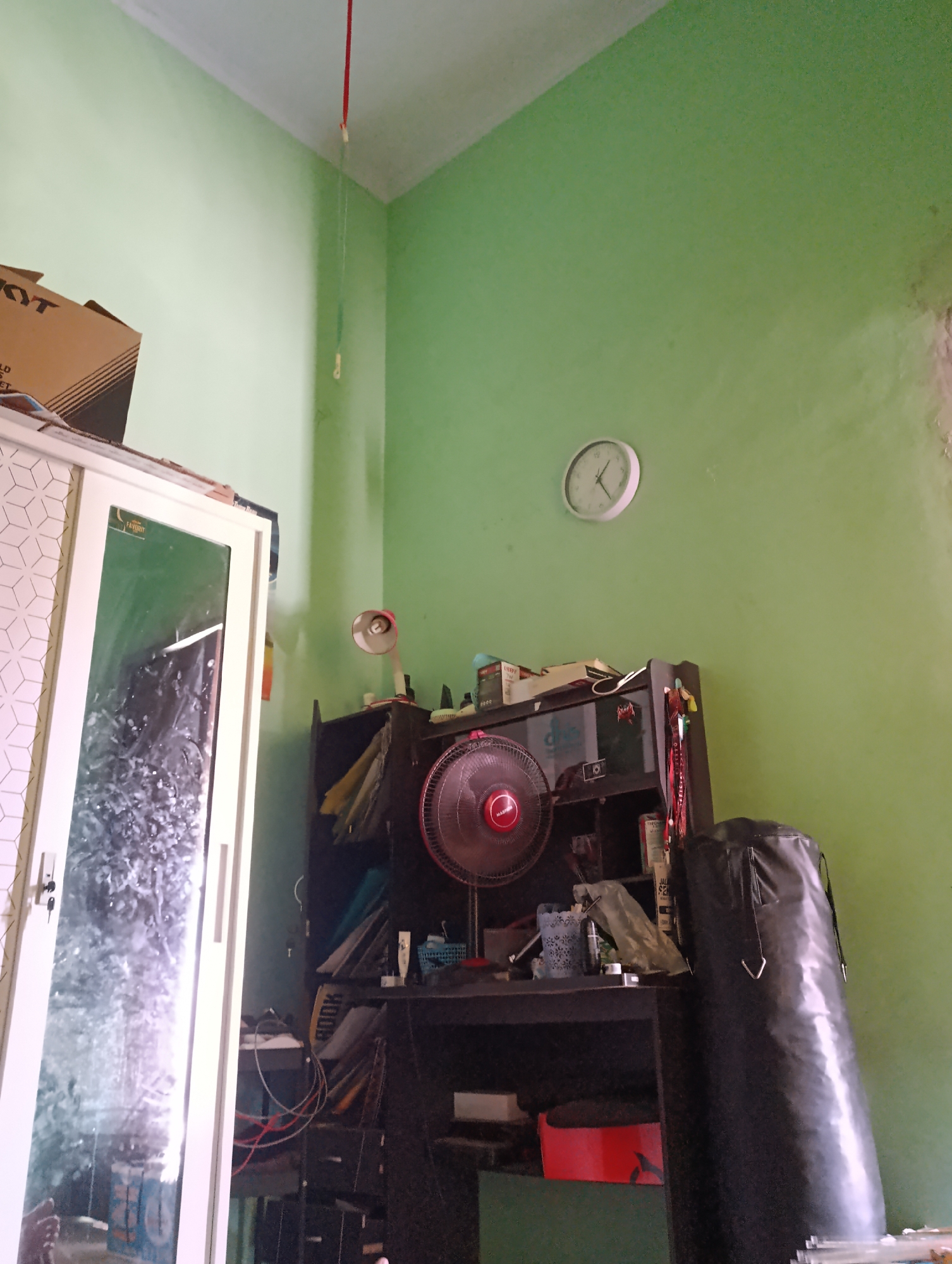 This image depicts an indoor scene within a room, potentially a bedroom or study, located in Jombor, Indonesia. The walls of the room are painted a light green, and the ceiling is white. A large, white wardrobe with a mirrored door is positioned on the left side of the frame. Above the wardrobe, a cardboard box with "KYT" printed on it is visible.
In the center of the image, a dark wooden desk is situated against the wall. On the desk, a red-bladed fan is prominently displayed, along with various items including what appears to be a lamp with a red shade, some bottles, and a small black camera. Behind the desk, a black shelving unit holds several items, including books, folders, and other assorted objects.
To the right of the desk, a large, black punching bag stands upright, partially obscuring the wall. A round, white clock with black hands is mounted on the green wall above the desk, indicating a time close to 11:00. A thin red wire, possibly for a light fixture, hangs from the ceiling in the upper left portion of the image. The overall impression is of a lived-in space with personal belongings.