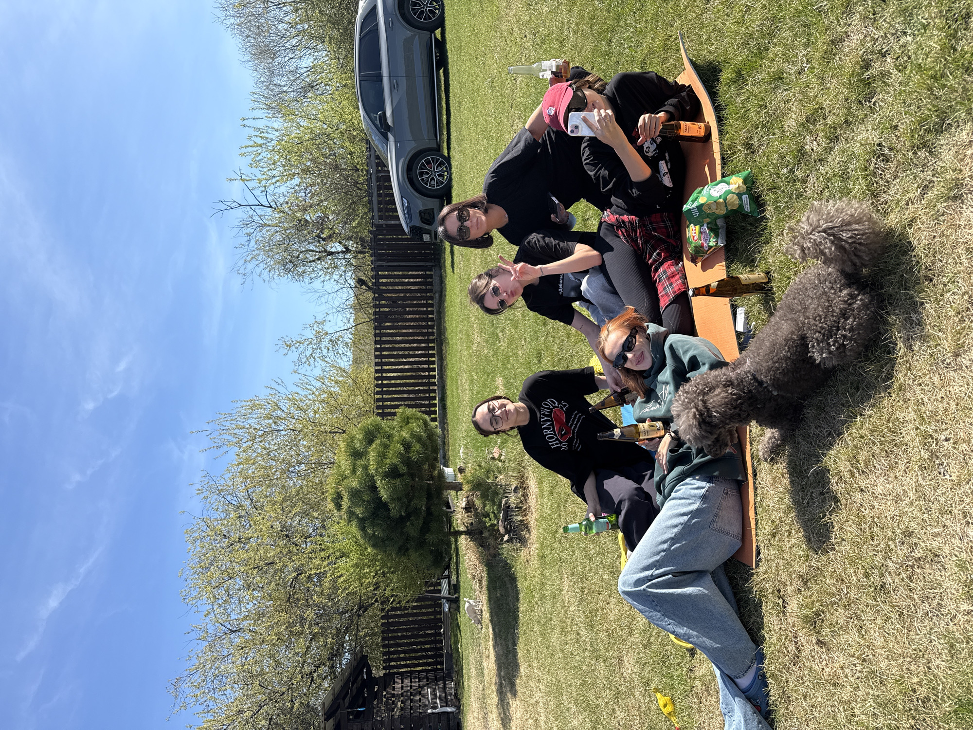 A group of four women and a grey poodle are in a grassy yard on a sunny day in Emel'yanovo, Russia. The women are sitting or lying on a sheet of brown material, with a bag of chips and several beer bottles nearby. One of the women is holding a cell phone, possibly taking a picture. A grey SUV is parked on the grass behind them, near a wooden fence. There are trees with sparse foliage in the background, and the sky is blue with light clouds. A small wooden structure is visible in the lower left corner of the image.