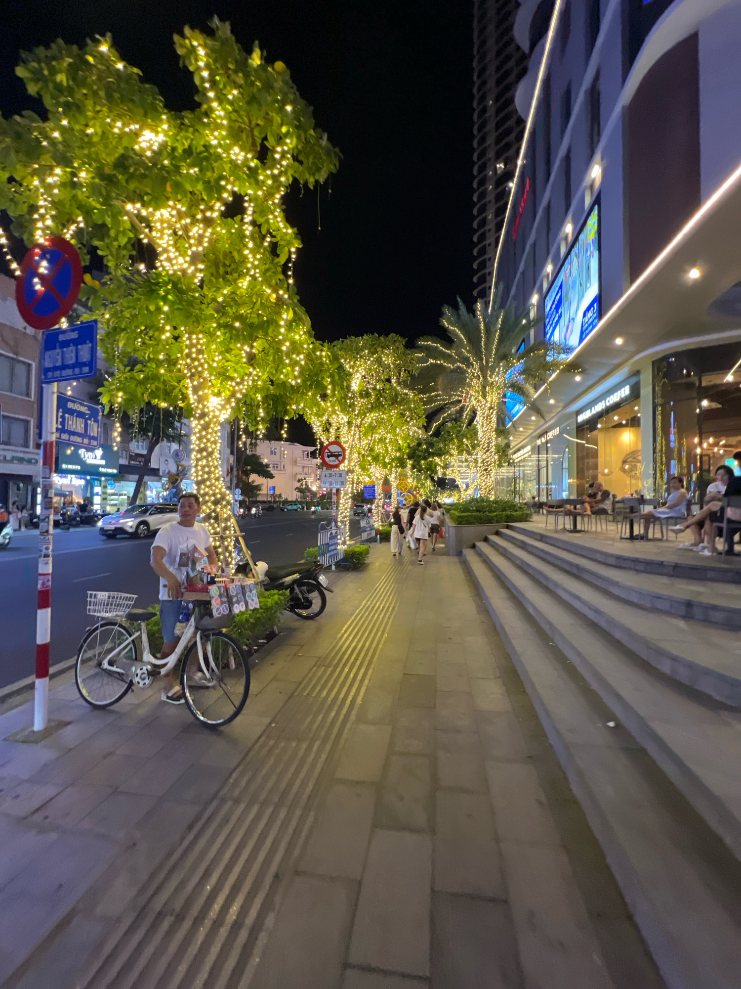 This image captures a street scene at night in Nha Trang, Vietnam, likely along a promenade or a busy commercial street. The foreground is dominated by a white bicycle parked on a sidewalk paved with grey flagstones and textured tactile paving. A man in a white t-shirt and blue shorts stands beside the bicycle, which has a basket filled with various items, possibly snacks or small goods for sale. The bicycle is decorated with colorful plastic rings and some packets. To the right of the sidewalk, a modern building with large glass windows and an illuminated facade rises up, housing establishments like Highland Coffee. Rows of outdoor seating are visible in front of the building, with several patrons seated at tables.
The street is lined with trees adorned with numerous warm-toned fairy lights, creating a festive and inviting atmosphere. In the background, traffic can be seen on the road, with cars and a motorcycle visible. Several street signs and traffic signals are also present. The overall lighting suggests it is evening or nighttime, with the artificial lights from the building, trees, and streetlamps illuminating the scene. The presence of people walking along the sidewalk and seated at the cafe indicates a lively urban environment.