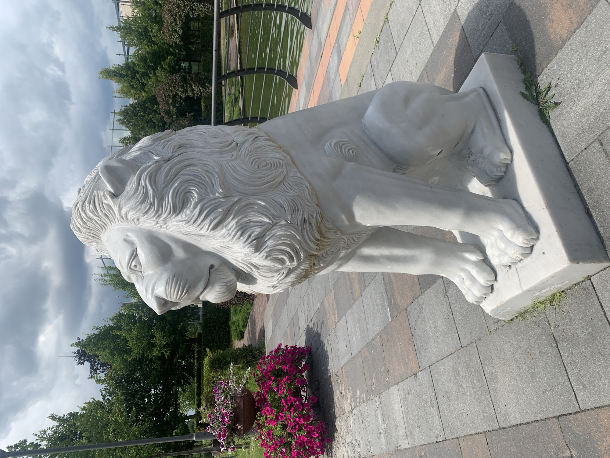 A white stone lion sculpture is positioned on a paved walkway next to a body of water. The sculpture is in a seated pose, facing left, with its mane elaborately carved. In the background, lush green trees and a cloudy sky are visible. To the left of the sculpture, a large pot overflows with bright pink flowers. The walkway is constructed of square, gray and brown paving stones. A decorative metal fence separates the walkway from the water. The location is identified as Bucha, Ukraine.