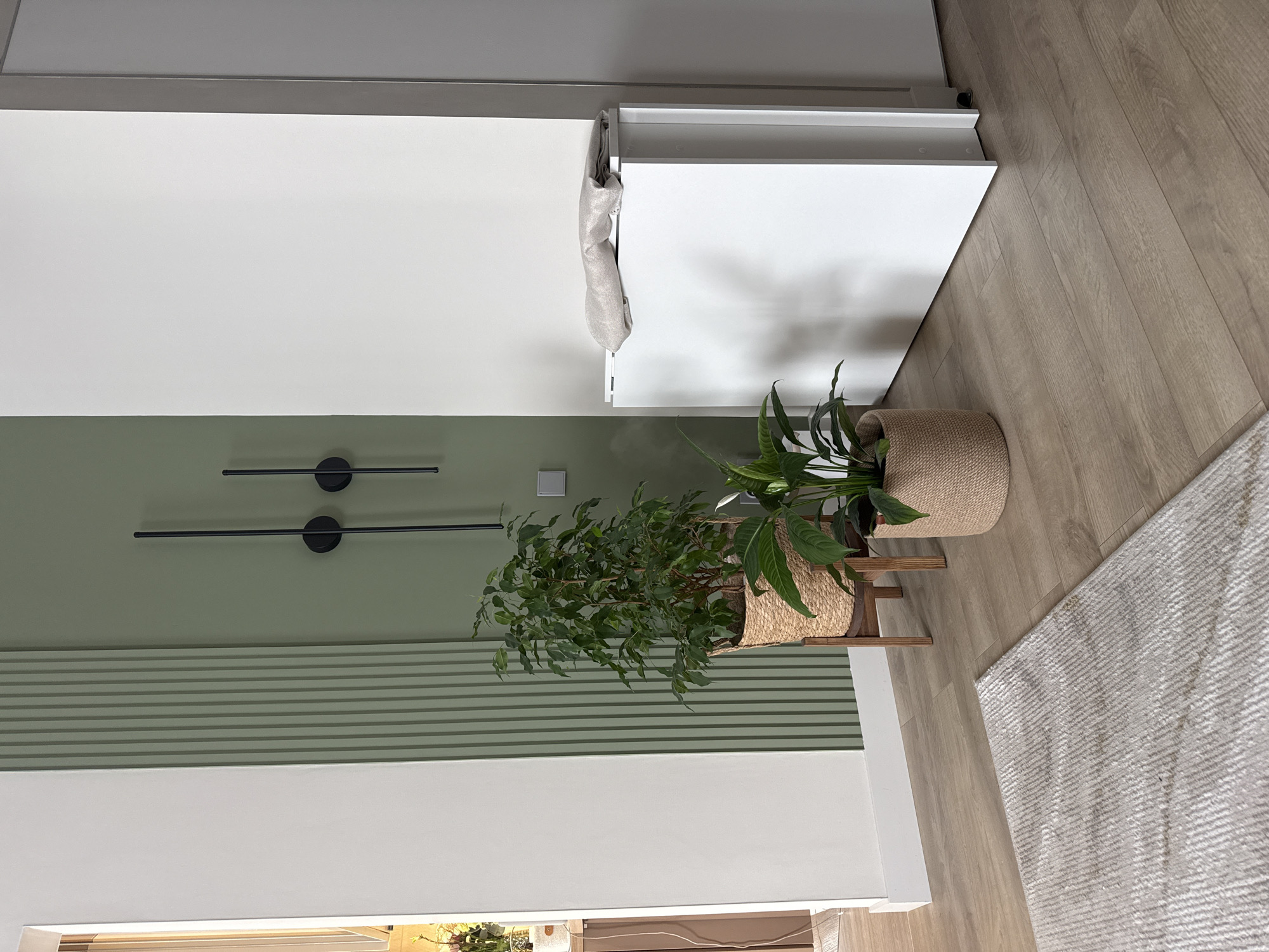 The image shows an interior with neutral tones. A green wall with horizontal striped paneling is contrasted by a white cabinet and door. Two houseplants in woven baskets are placed on the floor, with a light-colored rug partially visible. A black wall clock with horizontal arms is present on the green wall. The flooring is a light wood-grain laminate. The image suggests a modern or contemporary style. The location in Nizhnevartovsk, Russia, cannot be determined from this image.