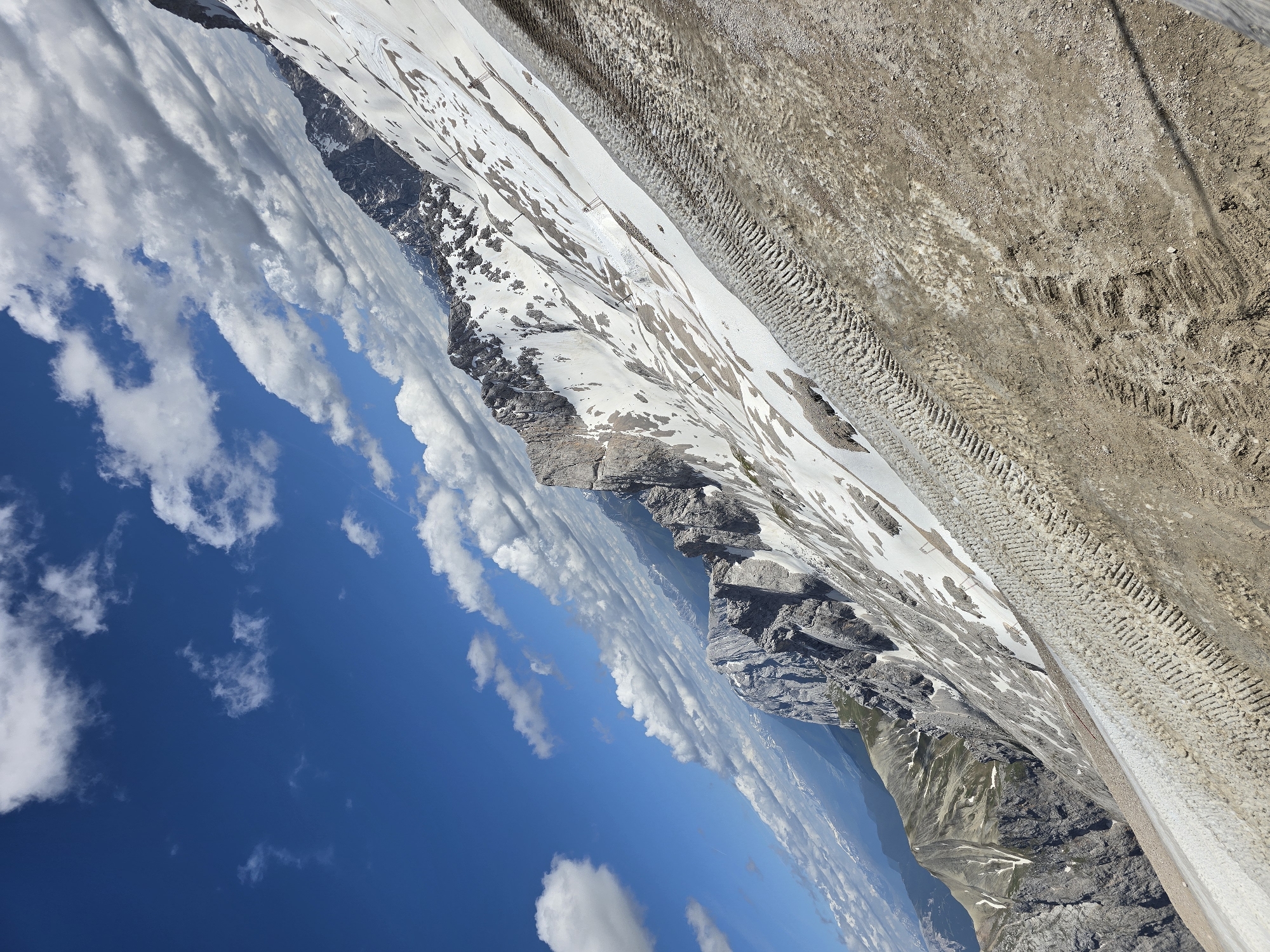 The image depicts a steep, snow-covered mountainside under a bright blue sky with scattered white clouds. The terrain transitions from rocky peaks and patches of green vegetation in the distance to a vast expanse of snow with visible tire tracks on a dirt path in the foreground. The snow is not continuous and shows patches of exposed rock and dirt, suggesting a melting or receding snowline. The lighting indicates it is daytime. The scene is likely a mountain landscape in the vicinity of Garmisch-Partenkirchen, Germany, possibly on a hiking trail or ski slope during a warmer season. There are no people or man-made structures visible in detail, though the tire tracks suggest recent vehicle activity.