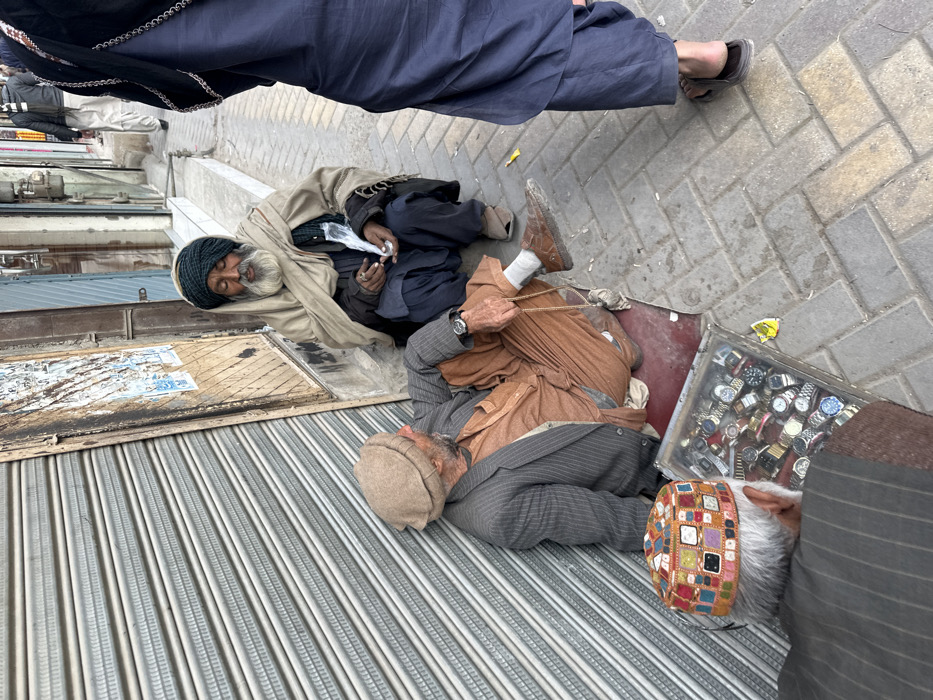 Three older men are lying on the ground next to each other. One wears a turban and a blanket and is holding paper or money. Another is wearing a blazer and cap with a watch on his left wrist. The third man is in a striped shirt and wearing a colorful cap. He is lying next to a box full of watches. A fourth person is wearing blue pants and sandals and is walking through the frame.