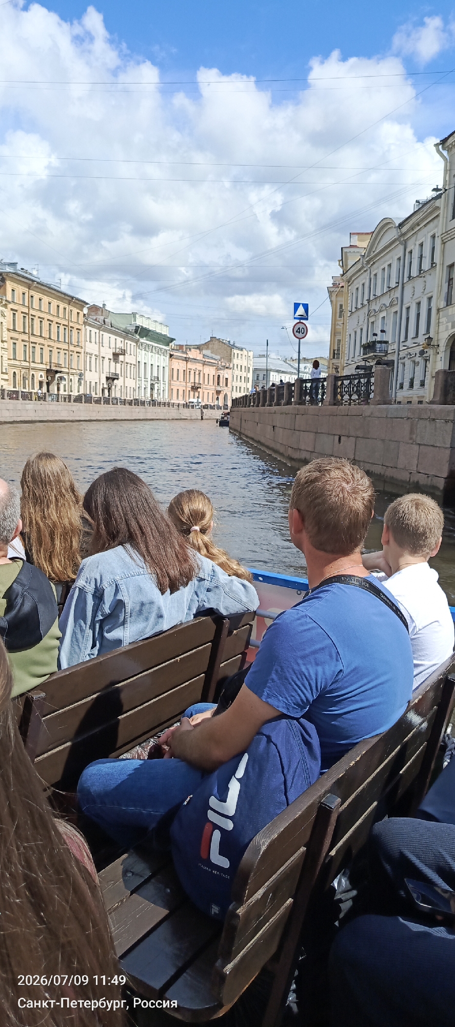 The image shows a canal boat tour in Saint Petersburg, Russia. Several people are seated on the boat, facing away from the camera. They appear to be enjoying a sunny day, with colorful buildings lining the canal in the background. A speed limit sign is visible on the right side of the canal. The water in the canal is calm, reflecting the blue sky and white clouds. The date and location are stamped in the bottom left corner: "2026/07/09 11:49 Санкт-Петербург, Россия".