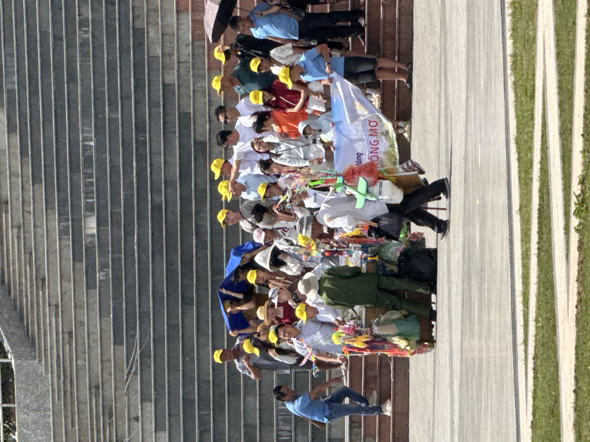 A group of people are gathered on wide stone stairs, likely in a public space in Da Lat, Vietnam. The stairs are arranged in broad, shallow steps with a concrete surface, leading upwards. In the foreground, a man in a blue shirt and jeans is descending the stairs, while to his right, a cart laden with colorful merchandise, including what appear to be toys, bags, and fabrics, is positioned. Behind the cart, a person in a green uniform stands. A large white banner with Vietnamese text, possibly advertising a product or event, is held up by several individuals. The crowd is diverse, with many wearing yellow baseball caps and casual clothing. Some are looking at the cart, others are interacting with each other, and some are shielded from the sun by an umbrella. The atmosphere appears to be that of a marketplace or a gathering point for tourists or locals. The background shows more of the stone stairs and hints of vegetation.