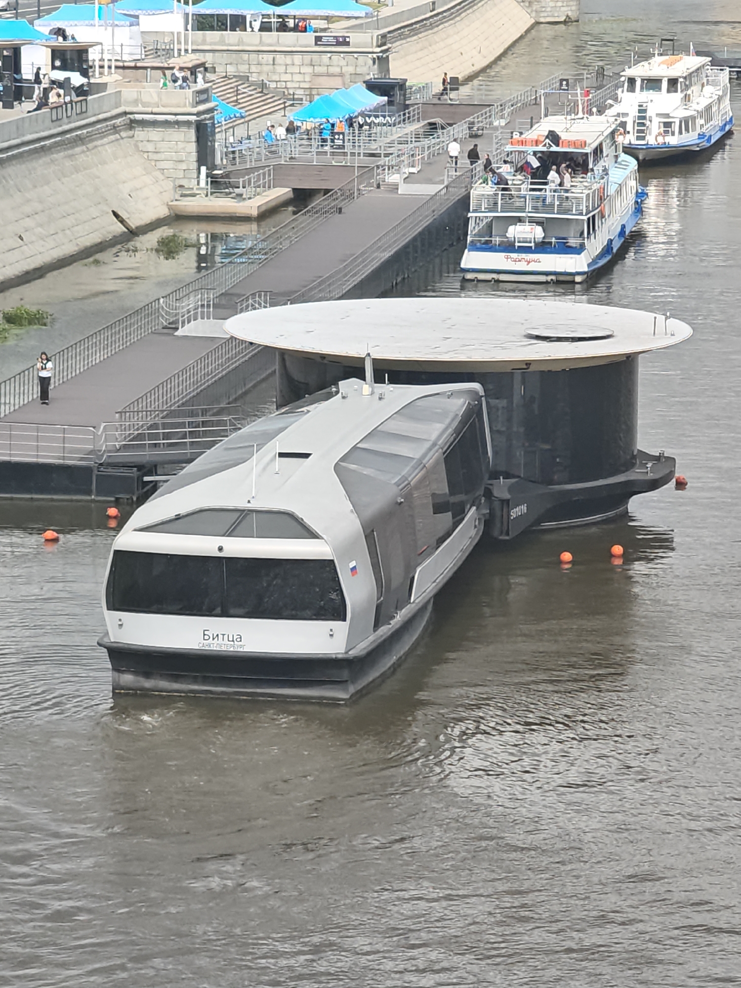 An aerial, high-angle shot shows a modern, grey and white watercraft docked at a circular, gray platform on a body of water. The watercraft has large, tinted windows and the word "Битца" and "САНКТ-ПЕТЕРБУРГ" written in white lettering on its stern, along with a Russian flag. Two orange buoys float in the water near the stern of the watercraft. In the background, a long, dark grey walkway extends from the platform towards a concrete embankment. Further in the background, two white passenger boats are docked. One boat has "Роксун" written on its stern in red lettering. Several people are visible on the boats and along the embankment, some of whom are under blue awnings. The scene appears to be a busy waterfront area in Moscow, Russia, likely a pier or promenade for river tours.