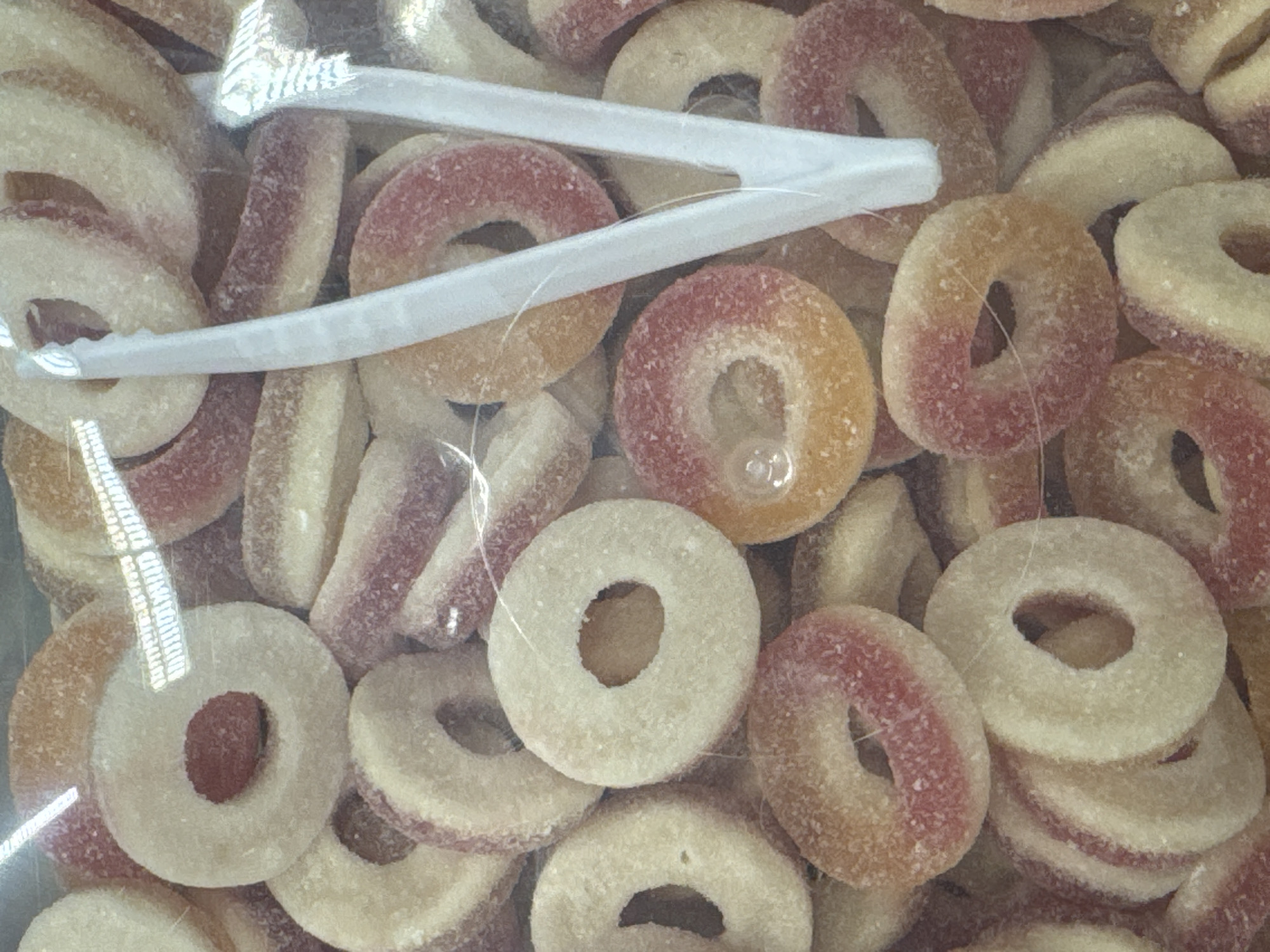 A close-up, top-down shot captures a pile of ring-shaped candies, possibly gummy rings, submerged in a clear plastic bag. The candies are predominantly a pale yellow or cream color, with distinct pink or reddish bands encircling their outer edges. A scattering of sugar crystals is visible on the surfaces of the candies, giving them a slightly frosted appearance.
Two white plastic tongs are partially visible, positioned in the upper-middle section of the frame, with their tips grasping some of the candies. The tongs appear to be either for serving or holding the candies within the bag. The lighting suggests an indoor setting, with some reflections on the plastic surface indicating overhead light sources. The overall impression is of a sweet treat assortment, likely a product for sale or consumption. No people, text, specific location cues, time of day, or weather conditions are discernible in the image.