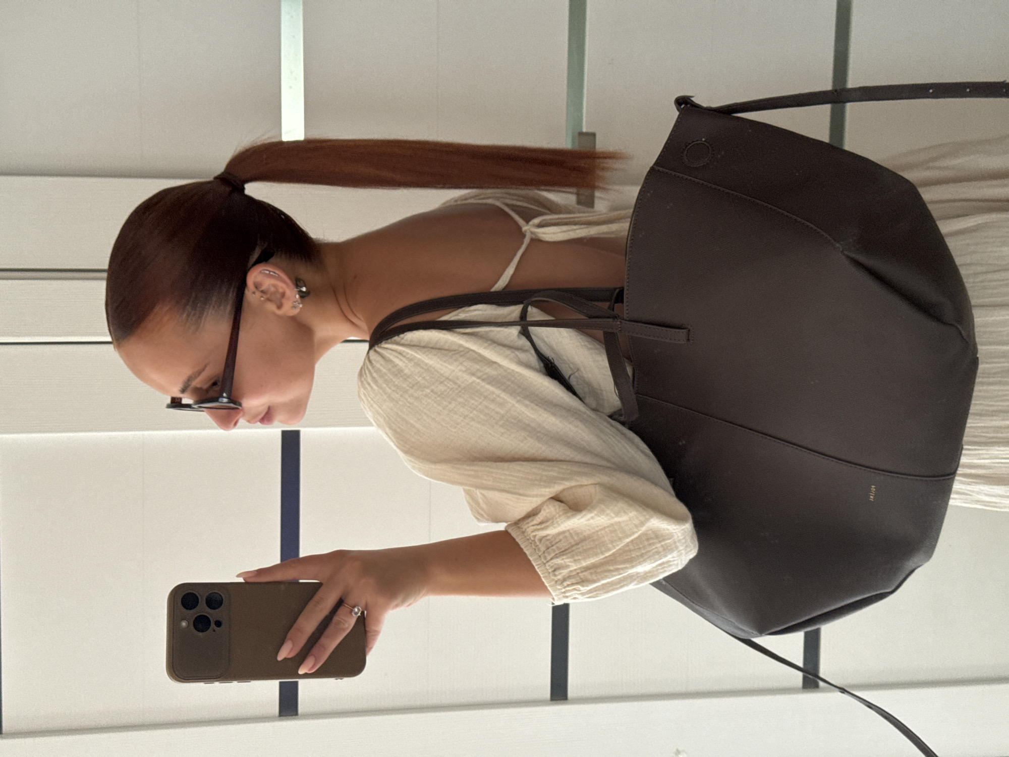 The image shows a woman taking a selfie in what appears to be an indoor setting with white walls and vertical slats of some kind. She has red-brown hair pulled back in a ponytail, and she's wearing sunglasses. She's wearing a light-colored, possibly linen or cotton, top with gathered sleeves. She is wearing earrings and a ring. She's holding a brown smartphone, likely taking the picture with it. A large, dark brown leather tote bag is slung over her shoulder. The bag has a shoulder strap.
There's no visible information to indicate this image was taken in Chaikovsky, Russia, or any specific location.