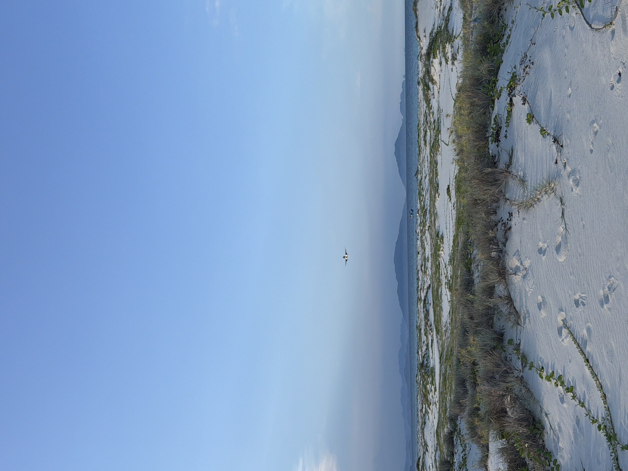 A serene beach scene unfolds under a clear blue sky. White sand dunes, sparsely covered with low-lying green vegetation, stretch across the foreground. Footprints are visible in the sand, suggesting recent human presence. In the middle ground, the calm, blue ocean meets a distant shoreline characterized by rolling, misty mountains. An airplane is captured in mid-flight, traversing the vast expanse of the sky. The scene evokes a sense of tranquility and natural beauty, likely in a coastal area like Bac Cam Ranh Ward, Vietnam, given the characteristic landscape.
