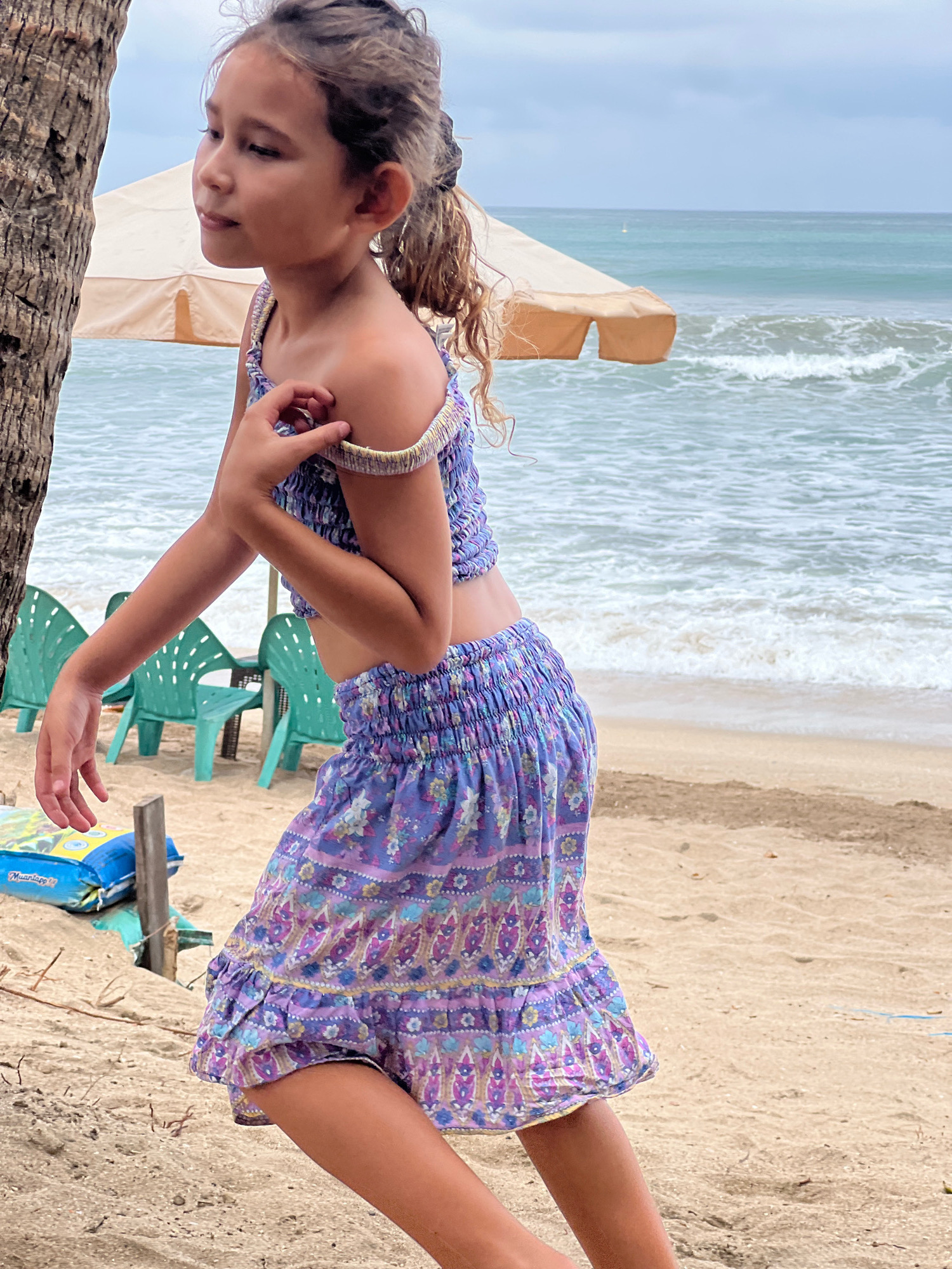 A young girl, appearing to be of Southeast Asian descent, stands on a sandy beach in Kuta, Indonesia, with the ocean and sky visible in the background. She is wearing a purple two-piece outfit with a floral pattern and ruffles. The top is a smocked crop top, and the skirt is a ruffled mini-skirt. Her hair is pulled back in a ponytail. Her left hand is near her shoulder, and her right arm is extended slightly. In the background, there are several teal plastic chairs, a beach umbrella, and a blue inflatable object partially obscured by sand and driftwood. The sky is overcast.