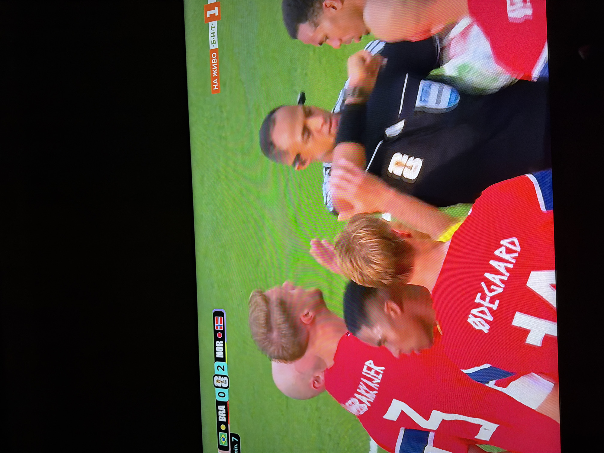 The image captures a scene from a soccer match, likely a television broadcast, displayed on a screen. At the bottom of the screen, a scoreboard indicates "BRA 0 2 NOR," with the flags of Brazil and Norway next to the respective country codes. To the right, text reads "НА ЖИВО Б-Н-Т 1," which translates to "LIVE BNT 1," suggesting the broadcast is live on Bulgaria's national television channel.
In the foreground, several individuals are visible. Two soccer players in red jerseys with white lettering are prominent. One player, on the left, has the name "BRAKKJER" and the number "7" on his jersey. The other player, closer to the center, has the name "ØDEGAARD" and the number "14" on his jersey. Both players appear to be of European descent.
A third individual, wearing a black referee's uniform with a FIFA logo on the chest and a badge on the arm, is positioned between the players. He has short dark hair and is looking downwards, possibly interacting with one of the players. His hands are near the players' arms, suggesting a moment of regulation or instruction.
Another player, partially obscured, is visible to the right of the referee, also appearing to be a player of color. The background is a green soccer field. The overall impression is a close-up shot of a moment during a soccer game, with a focus on the players and the referee. The location in Ognianovo, Bulgaria, is indicated by the television channel's presence in the broadcast.