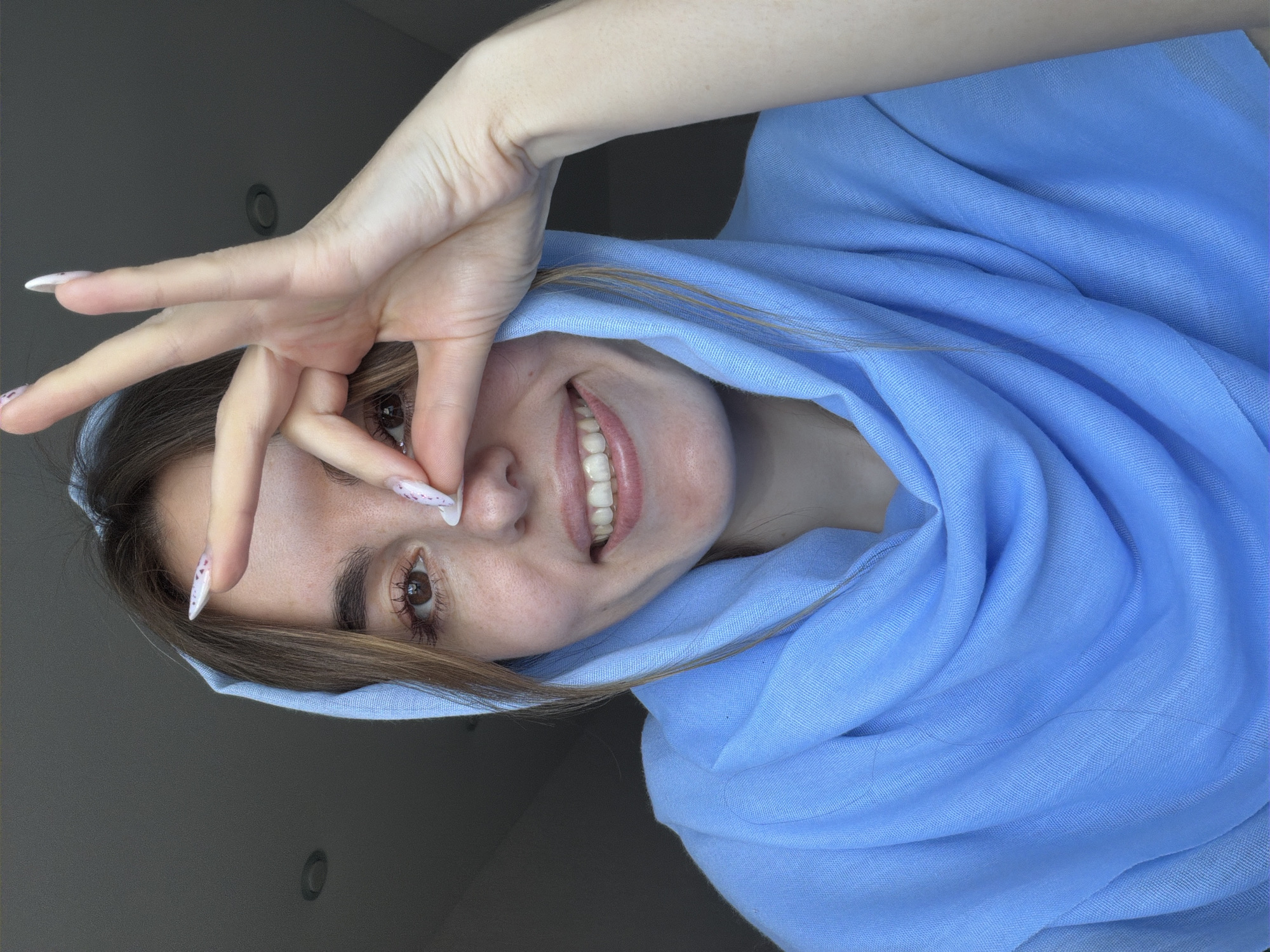 A person is indoors, wearing a light blue headscarf. They are smiling and making a gesture with their hand near their face. The background is a plain, light grey wall with two circular light fixtures. The overall lighting appears to be soft and diffused. There are no identifiable details to suggest a specific location in Sarapul, Russia.