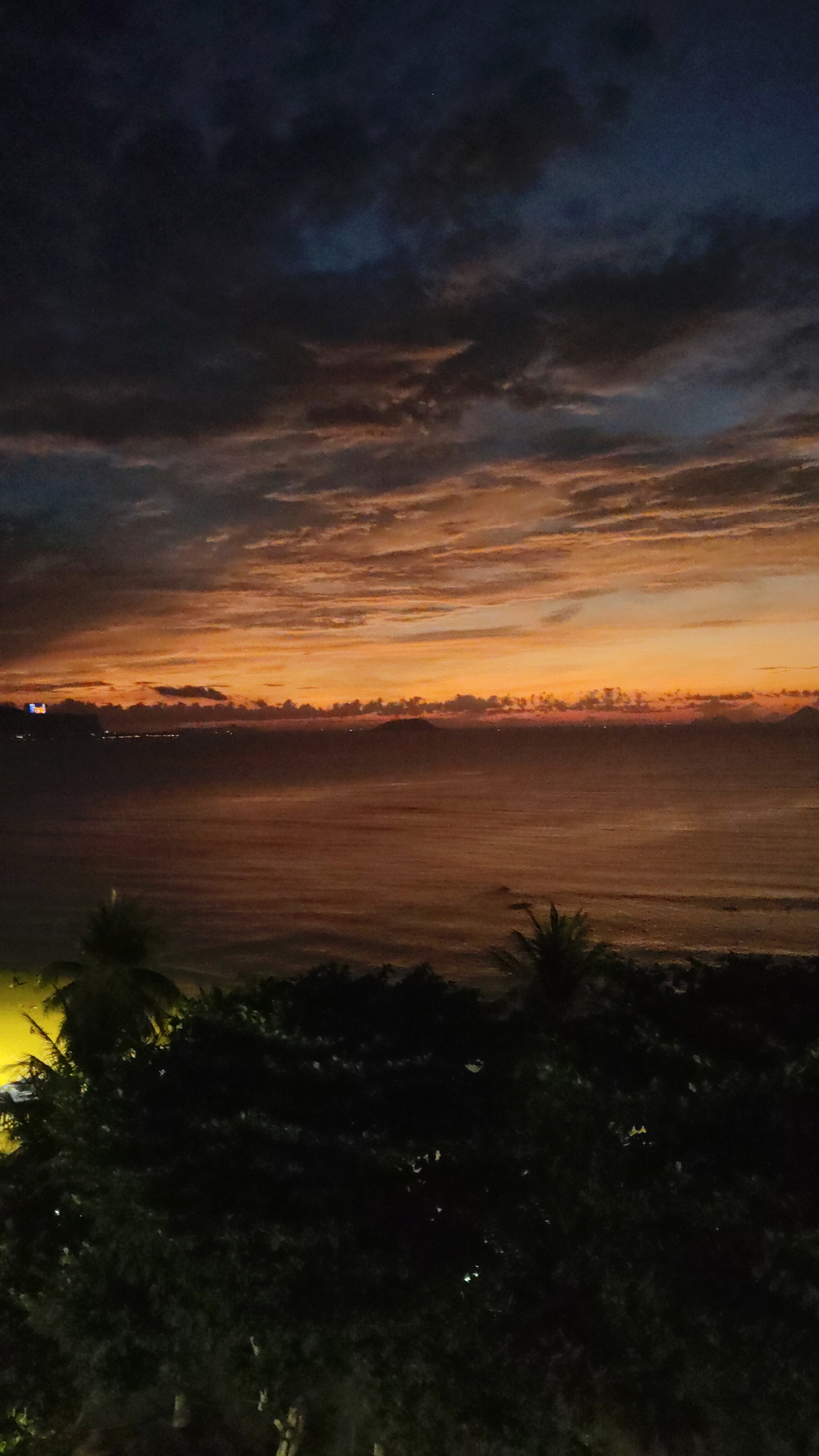 A nighttime view over the ocean in Bac Nha Trang Ward, Vietnam, reveals a vibrant sunset casting an orange glow on the clouds and water. In the foreground, the dark silhouette of trees, including palm trees, frames the scene. Distant lights are visible along the horizon, indicating a coastline or settlements. The sky above is filled with dark, cloudy formations, contrasting with the bright colors of the sunset. The water reflects the sky's colors, creating a unified color palette of orange, brown, and dark shades.