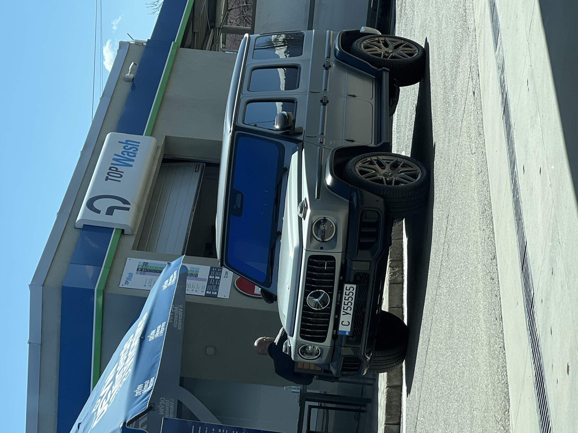 The image shows a silver Mercedes-Benz G-Class AMG parked in front of a "TOP Wash" car wash in Sofia, Bulgaria. The vehicle has a Bulgarian license plate, "C У55555 BC". A bald man in dark clothing is leaning against the front of the vehicle. The building has a blue and green color scheme with signage listing services and prices. The sky is clear, indicating a sunny day. The location appears to be an urban area near a street with a drainage system.