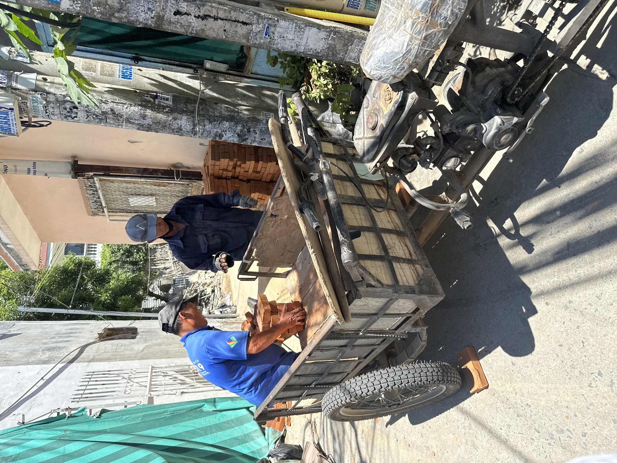 The image depicts an outdoor scene in what appears to be the city of Bac Nha Trang Ward, Vietnam. Two men are present. The man on the left, wearing a blue shirt and a grey baseball cap, is actively stacking bricks onto a wooden cart. His hands are gloved, and he is in the process of placing a brick. The man on the right, wearing a dark blue long-sleeved shirt and a grey cap, stands near the cart, observing or assisting. He also appears to be wearing gloves.
The cart itself is made of wood and metal, with a single spoked wheel visible on the left side. It contains stacks of bricks and some wooden planks. Attached to the rear of the cart is a motorized component, suggesting it might be a motorized cart or a modified bicycle.
The environment includes a narrow street or alleyway with buildings on either side. One building is painted a pale color, and another is a darker, possibly tiled structure. A green tarp is draped on a wall on the left side of the image. Electrical poles with wires and various stickers are visible in the background. Sunlight casts strong shadows across the ground, indicating it is daytime. The ground is paved with concrete or asphalt. A single brick is placed under the wheel of the cart. The overall scene suggests construction or material transport work.