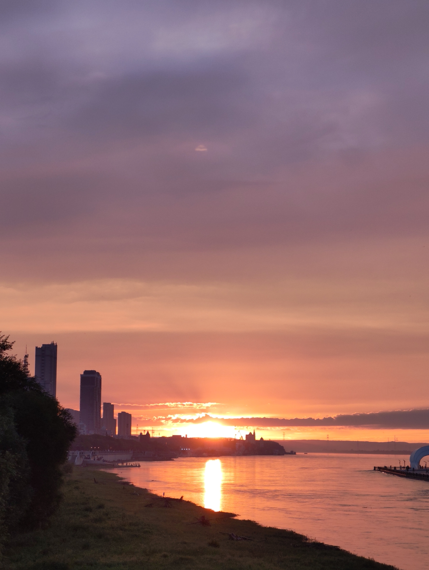 This image captures a sunset over a body of water in 幸福乡, China. A silhouetted cityscape with tall buildings lines the opposite bank of the river, bathed in the warm glow of the setting sun. The sky is a gradient of purples, pinks, and oranges, with scattered clouds adding texture. The sun's reflection creates a bright, shimmering path across the water. On the near bank, a grassy slope leads down to the water's edge. A pier or walkway extends into the water on the right side of the frame, with a distinctive arched structure. A few distant figures can be seen on the pier. The overall scene is one of natural beauty and urban presence.