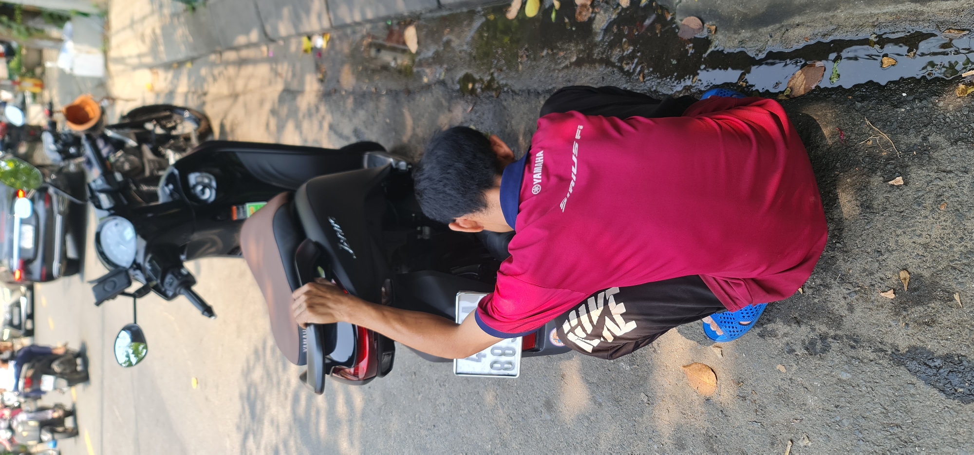 A man wearing a red Yamaha Sirius Fi shirt, black shorts, and blue sandals is crouching near a black Yamaha motorcycle parked on a street. He is working on the motorcycle, with his left hand on the seat and his right hand possibly adjusting something near the license plate. The license plate is white with black numbers. The street appears to be near a curb with some water and debris along the edge. Other motorcycles and vehicles are parked in the background. The location is likely in the city of Đà Nẵng, Vietnam, due to the presence of Yamaha branding and the visual style of vehicles.