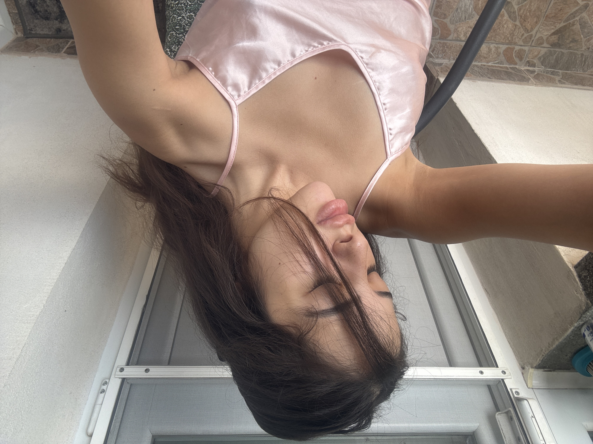 The image shows an upside-down close-up of a person wearing a pink satin camisole. The person's head is tilted back, with their eyes closed and their lips slightly parted. Dark hair falls around their face and shoulders. The camisole has thin spaghetti straps and a V-neckline. In the background, a white wall and a screened door are visible. To the right, a tiled surface and a dark object, possibly a chair, can be seen. The lighting suggests it might be daytime. The overall mood is relaxed and intimate.