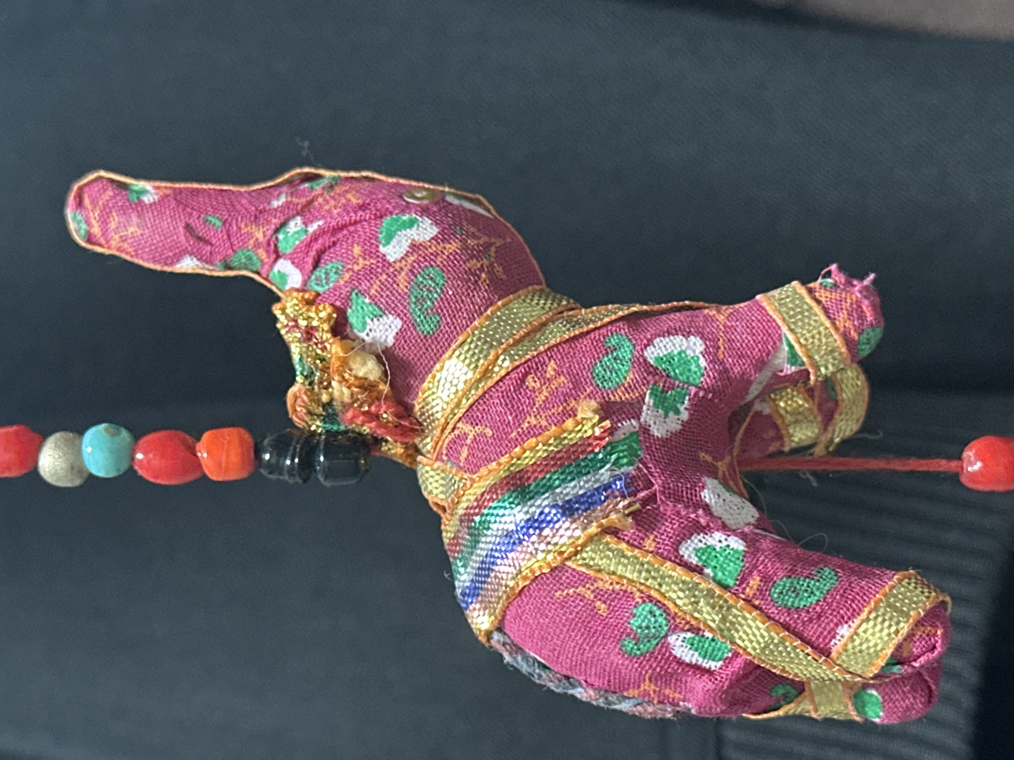 A close-up, vertical shot shows a small, pink, fabric elephant ornament hanging against a dark, out-of-focus background. The elephant is adorned with green and white floral patterns and decorated with gold trim. A strand of colorful beads, including red, turquoise, and black, hangs above the elephant. The overall scene is indoors, with no discernible elements indicating a specific location within Nevinnomyssk, Russia.